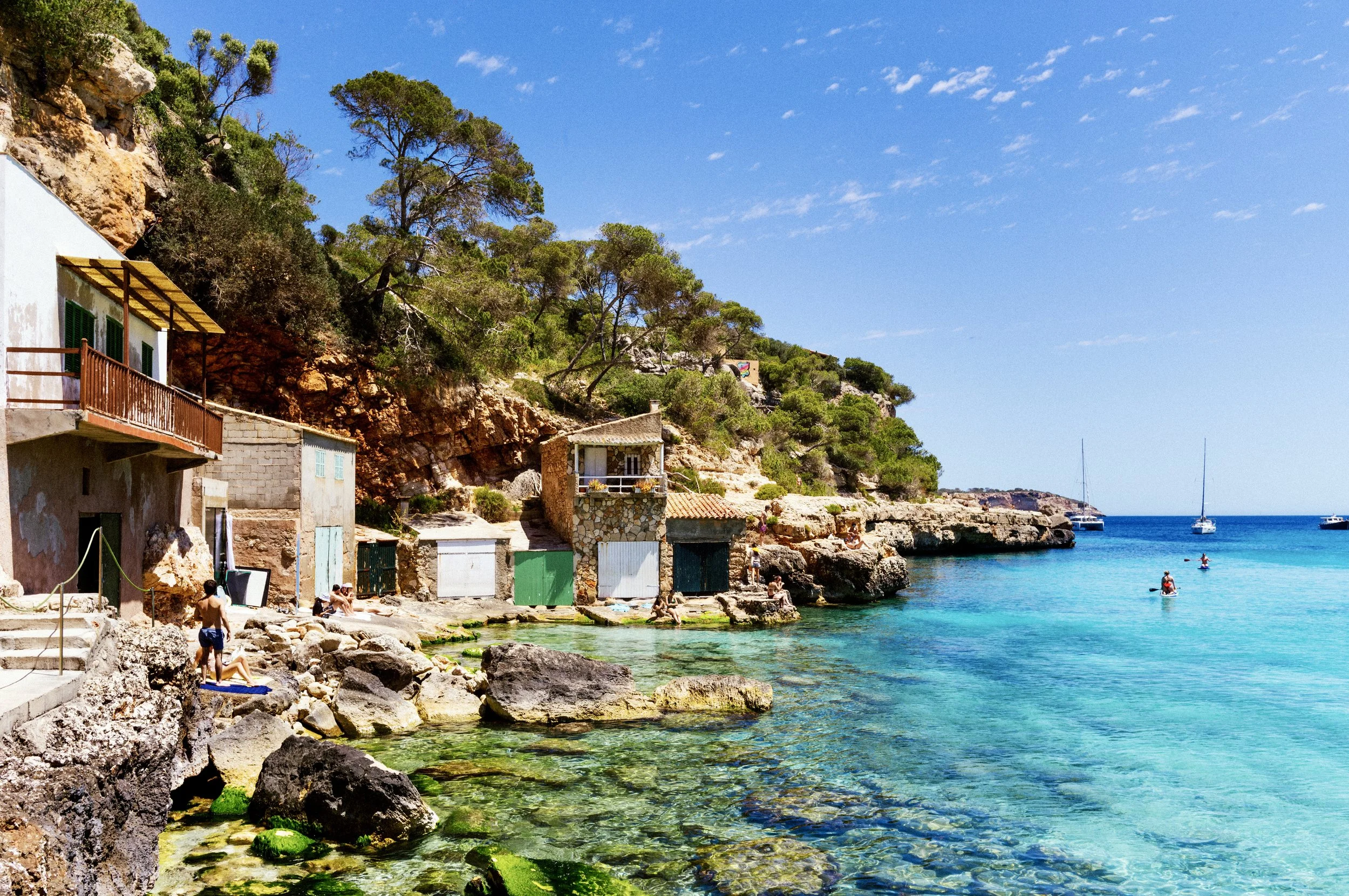 Sunbathers and traditional boat sheds beside crystal-clear water at a rocky cove in Mallorca