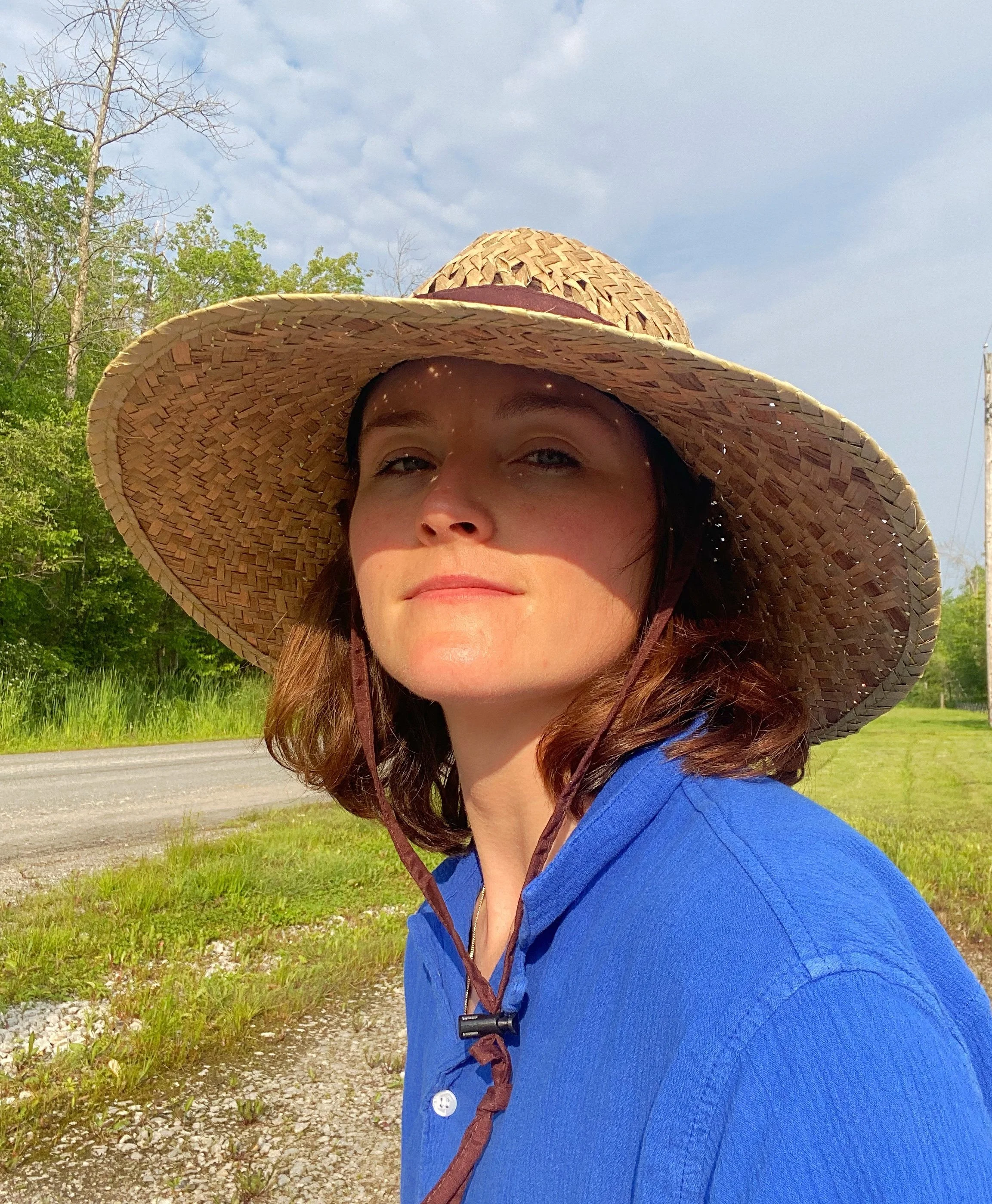 A person in a blue button down shirt wearing a straw hat looks at the camera outside.
