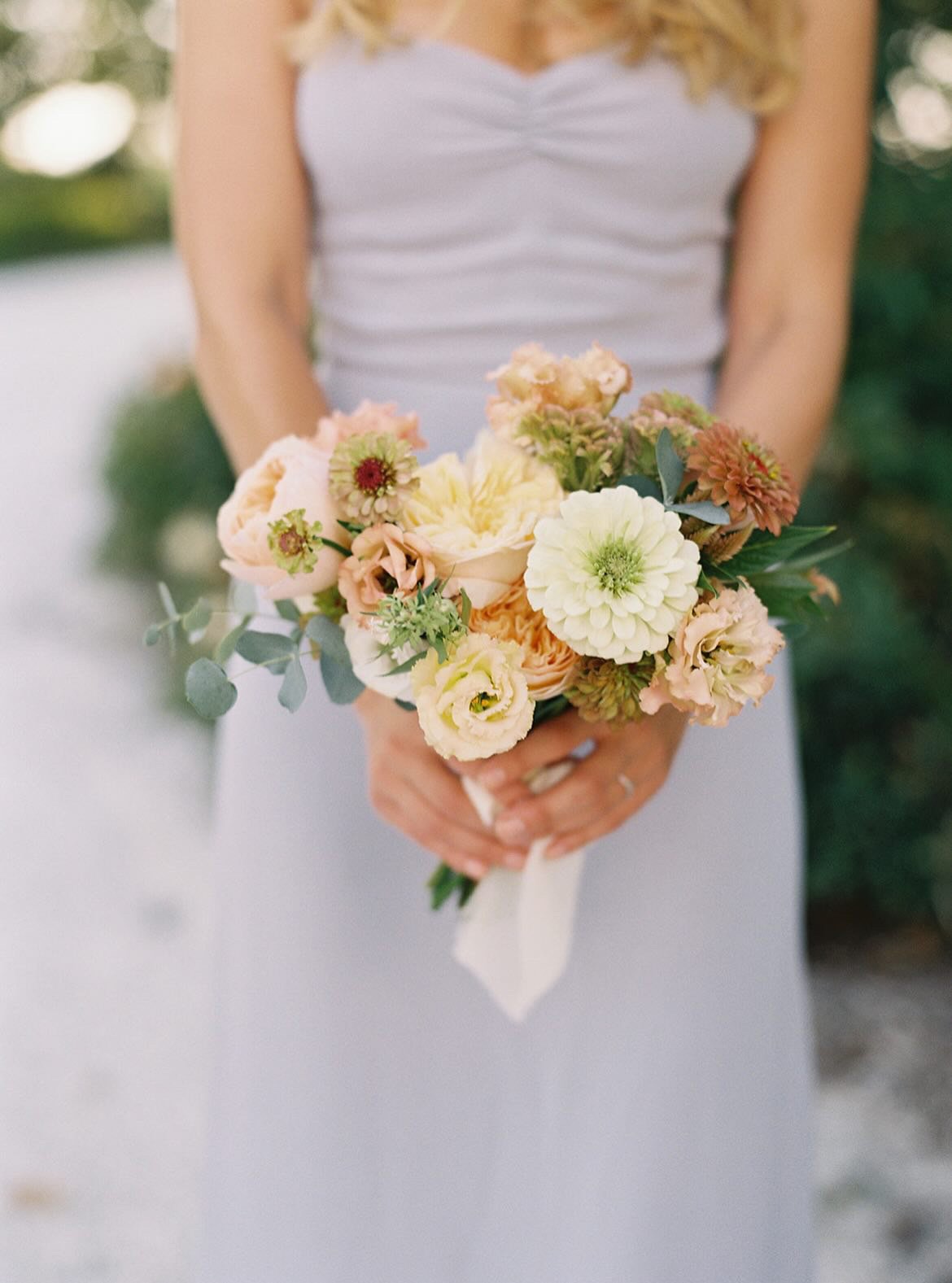 A woman in a light gray dress holding a bouquet of light-colored flowers including roses, dahlias, and greenery.