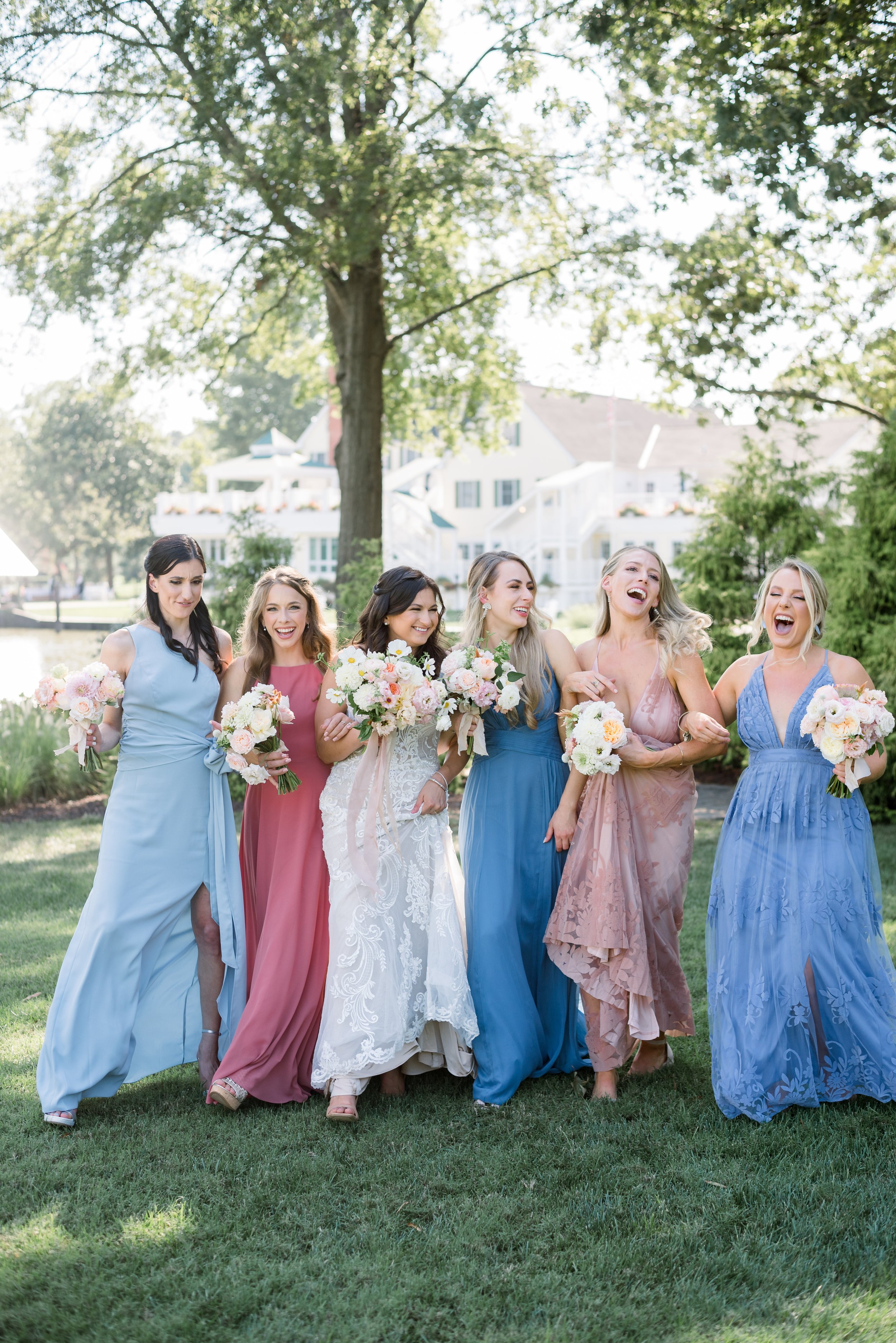 Group of six women in pastel dresses holding bouquets, walking on grass in a park with trees and white buildings in the background, enjoying a sunny day.