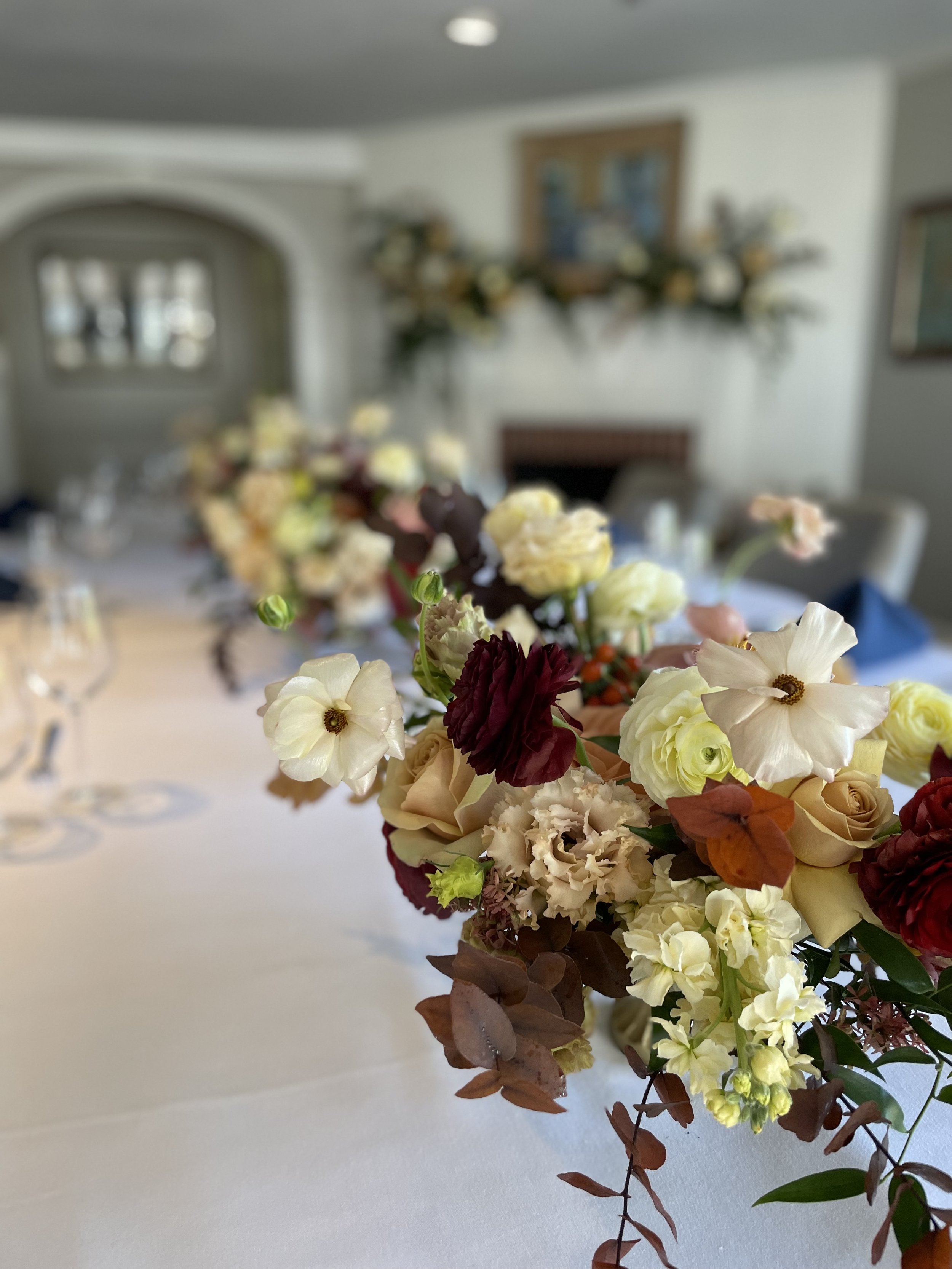 Close-up of a floral centerpiece with white, burgundy, and pale pink flowers on a white tablecloth, blurred background of a decorated dining room.