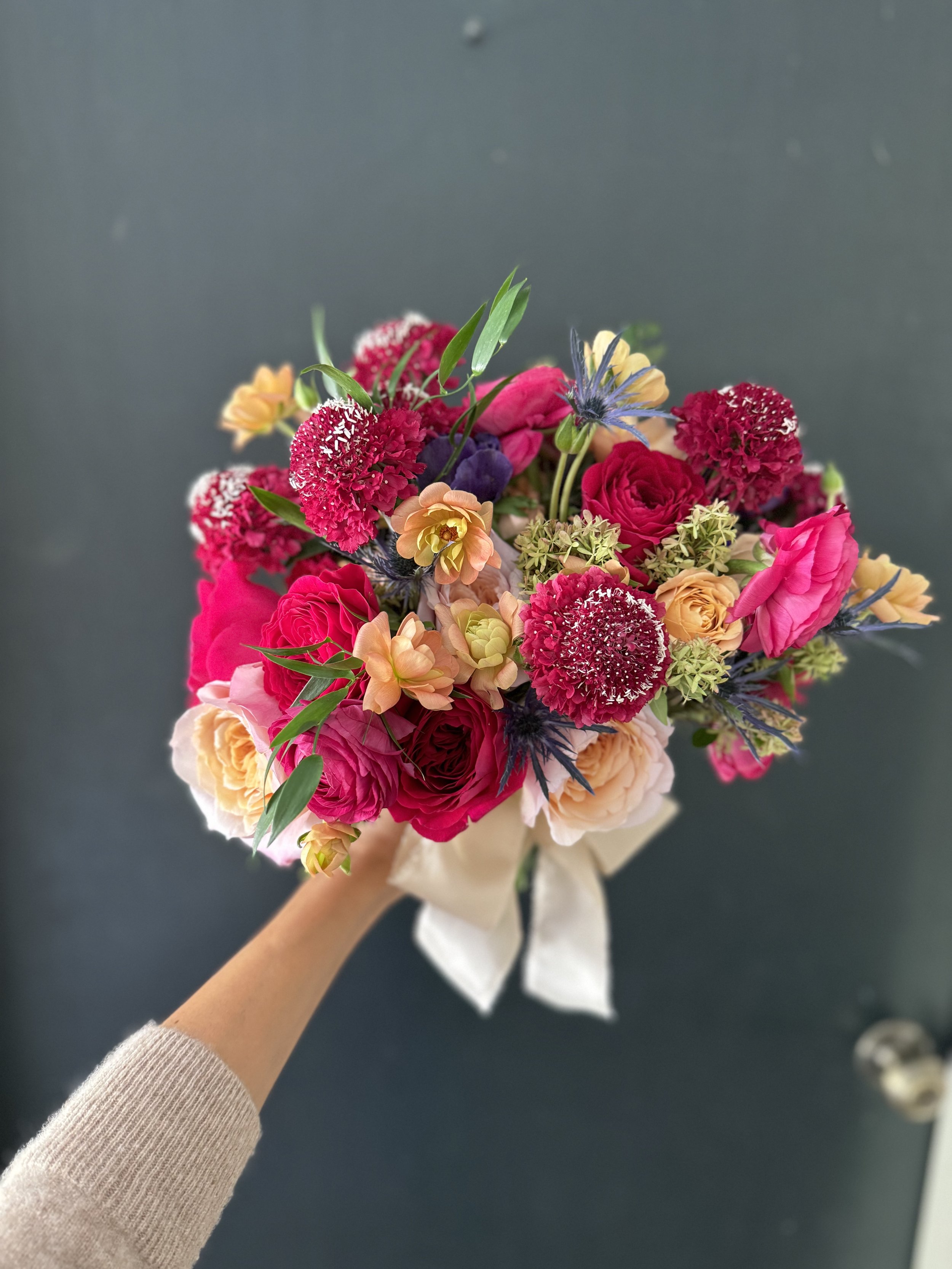 A colorful bouquet of various flowers including pink, red, yellow, and purple blossoms held with a white ribbon against a dark background.