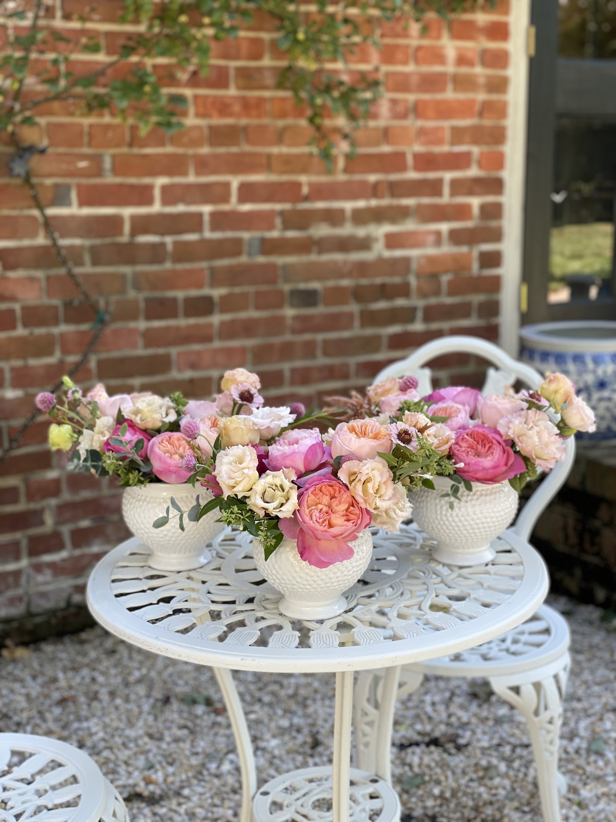 Three white ceramic vases with floral arrangements of pink, cream, and peach-colored roses and other flowers on a white metal outdoor table, with a brick wall and garden in the background