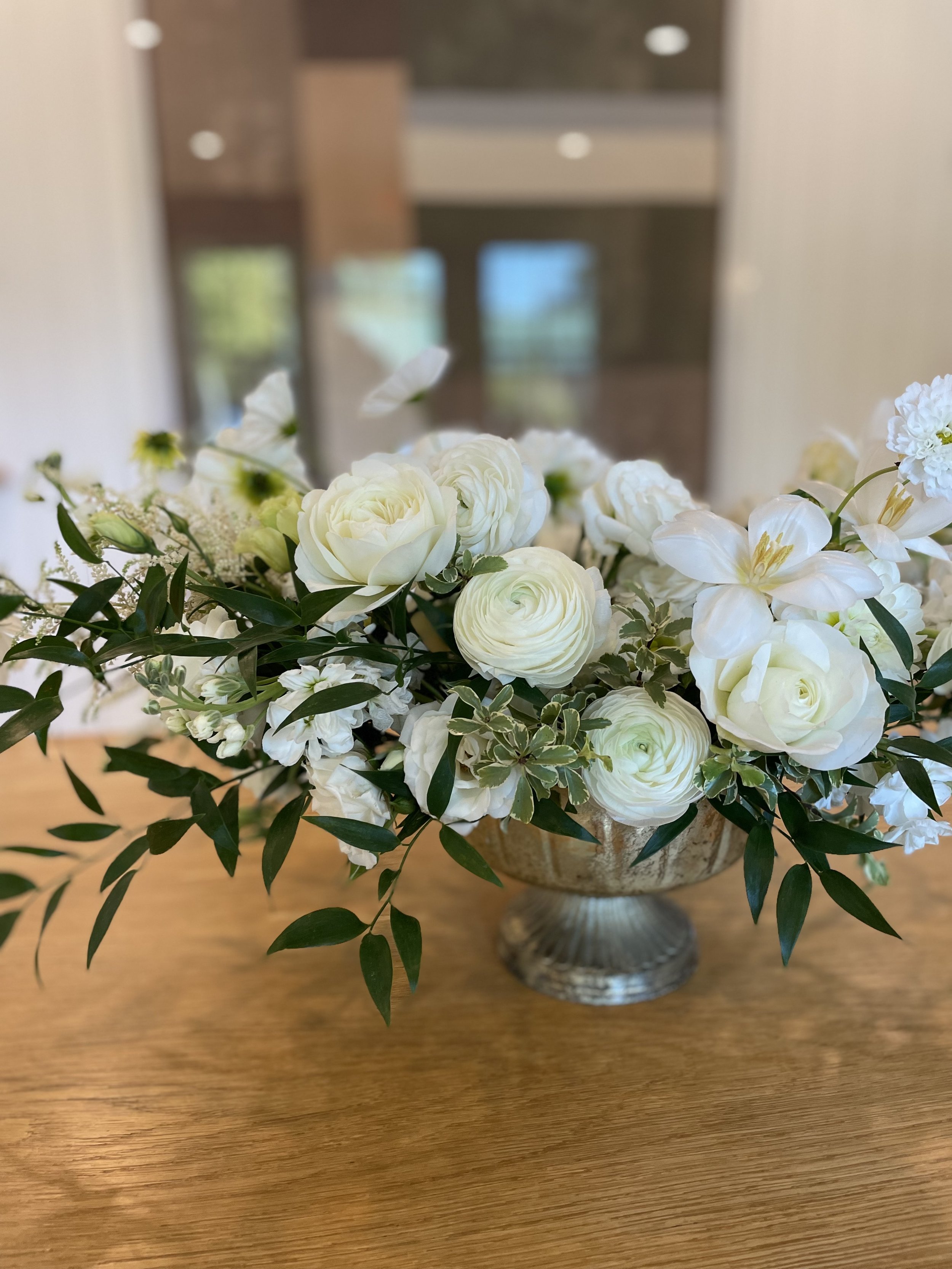 A bouquet of white flowers, including roses, ranunculus, and other blooms, arranged in a decorative silver bowl on a wooden table.