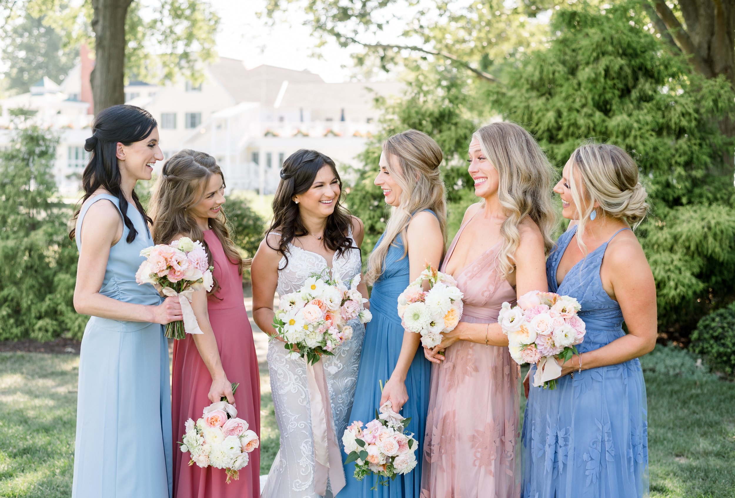 Group of seven women in colorful dresses holding bouquets, smiling and talking outdoors in a park during a wedding celebration.