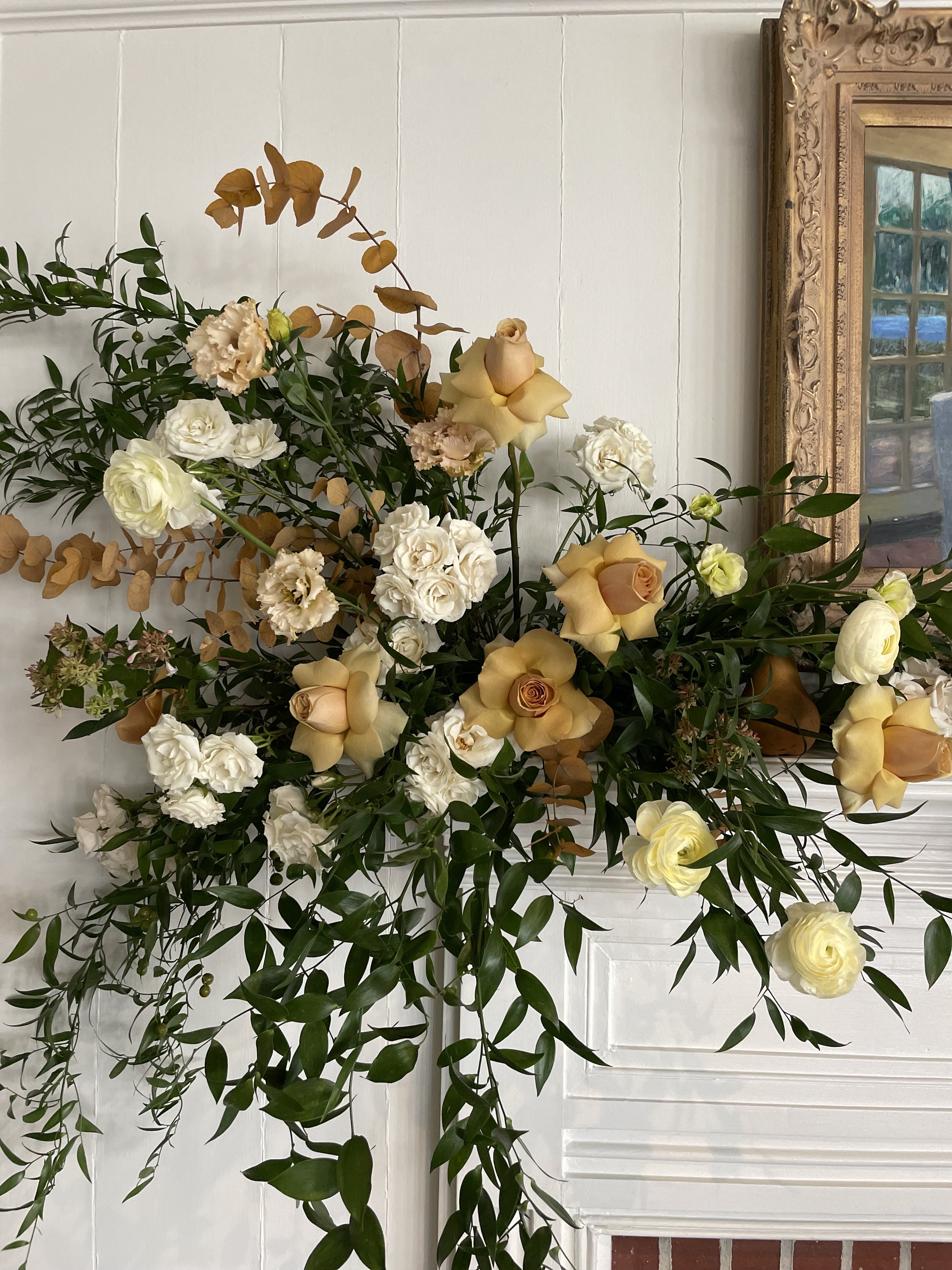 A large floral arrangement of white and cream-colored roses, lisianthus, and greenery, placed on a surface near a white wall and a decorative gold-framed mirror.