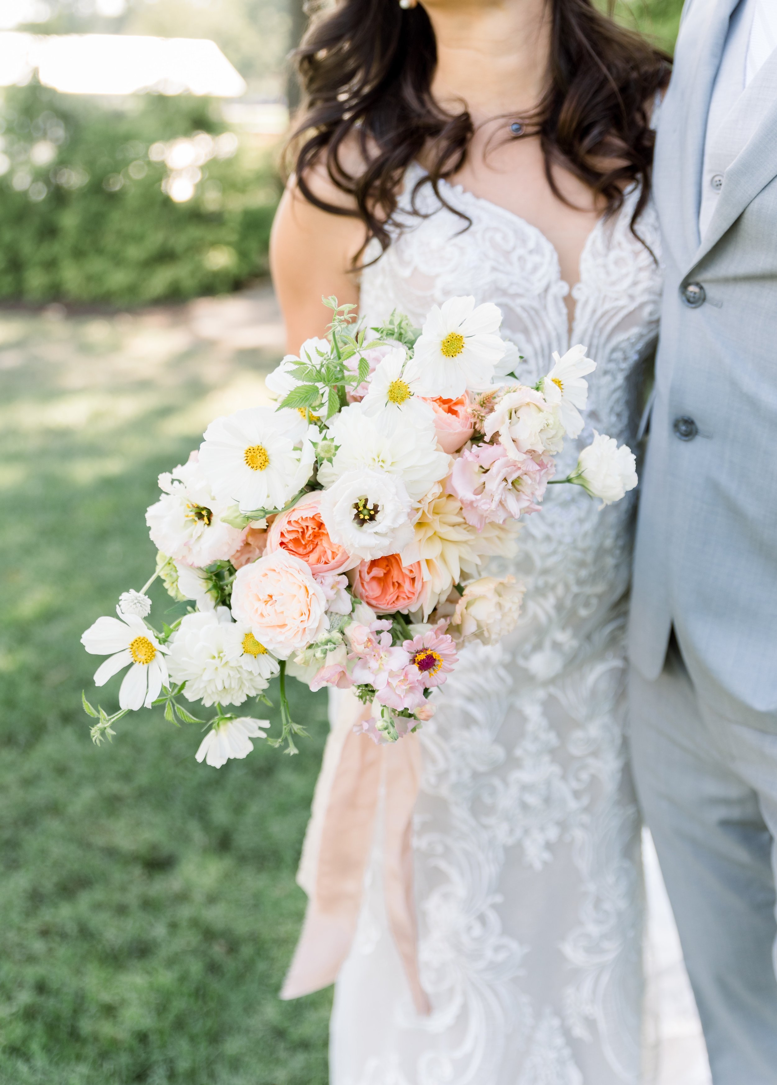 A bride and groom standing outdoors, with the bride holding a large bouquet of white, pink, and peach flowers. The bride is wearing a white lace dress and a silver necklace, and the groom is wearing a light gray suit.