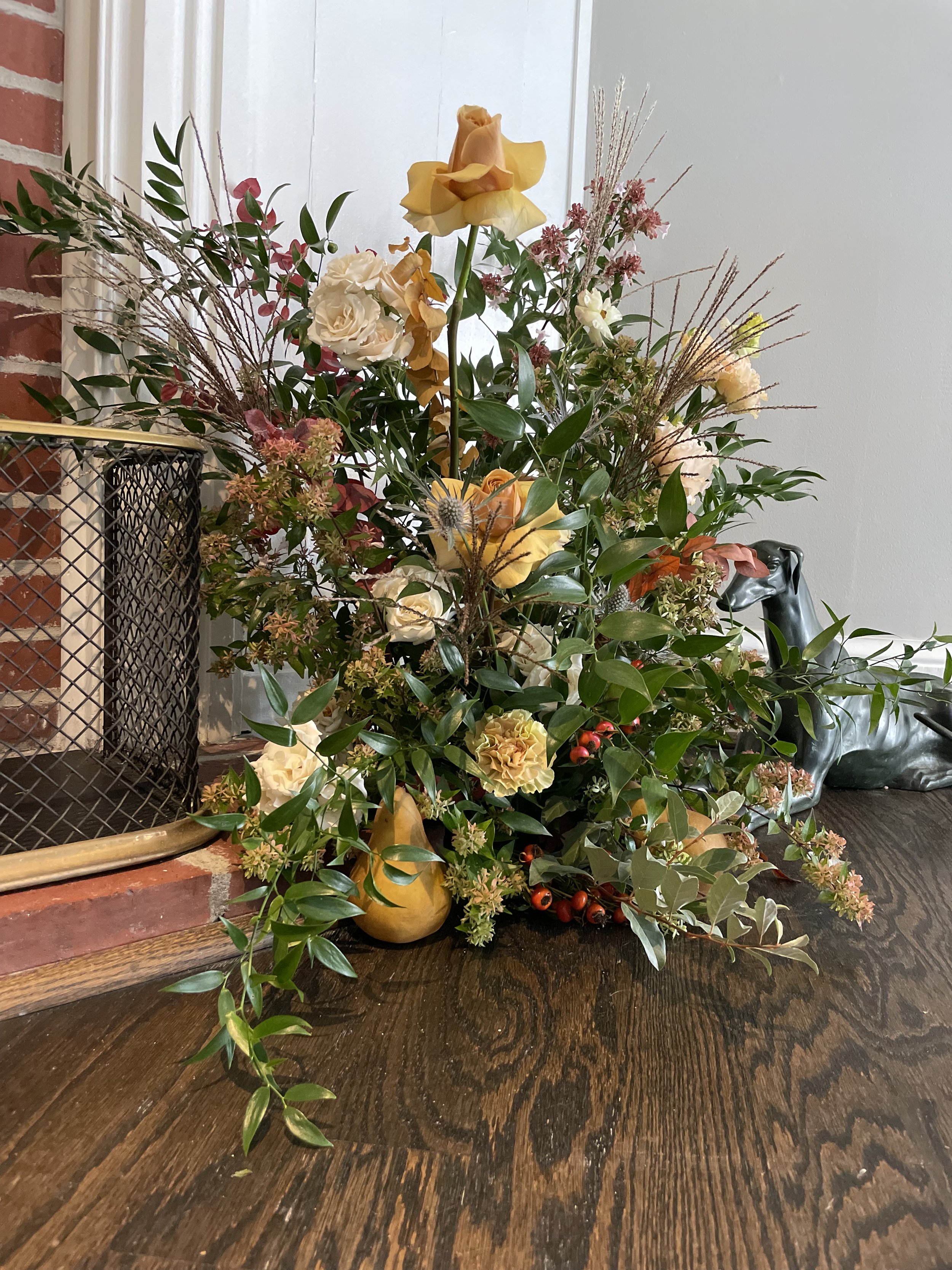 Decorative arrangement of yellow and white flowers with green leaves and red berries on a dark wooden table next to a brick fireplace.