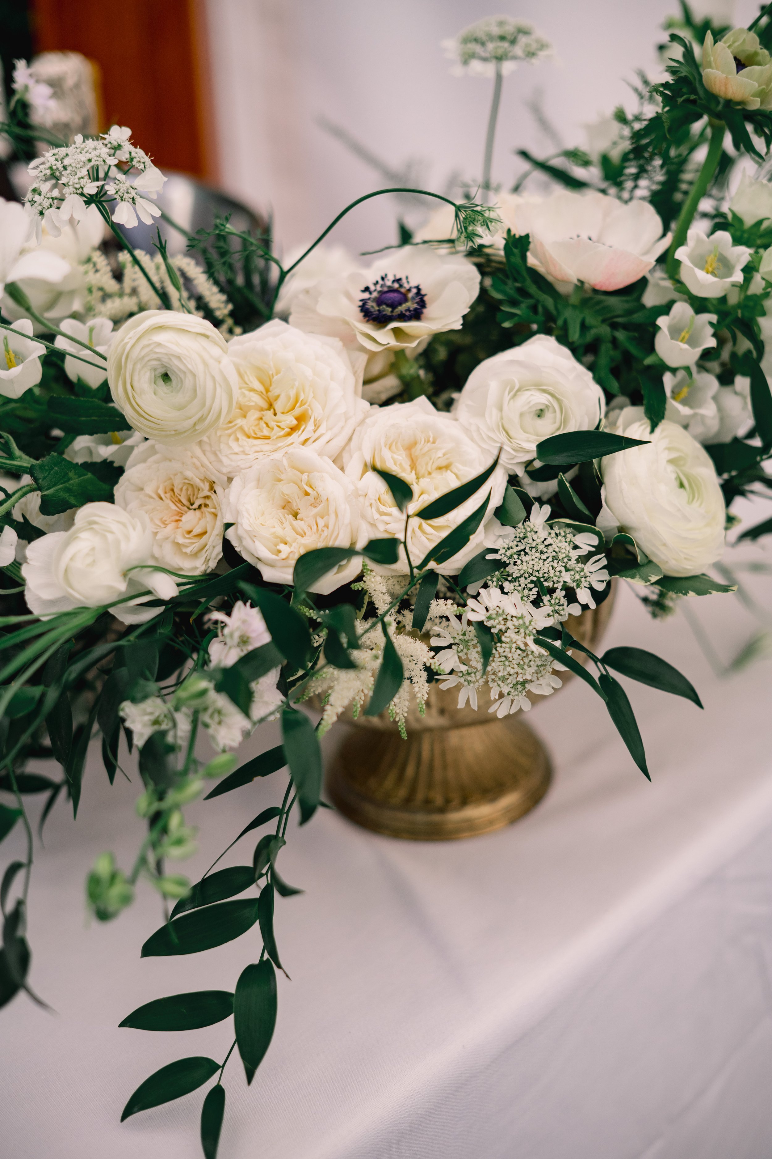 Elegant floral arrangement with white roses, ranunculus, anemones, and greenery in a gold vase on a white table.