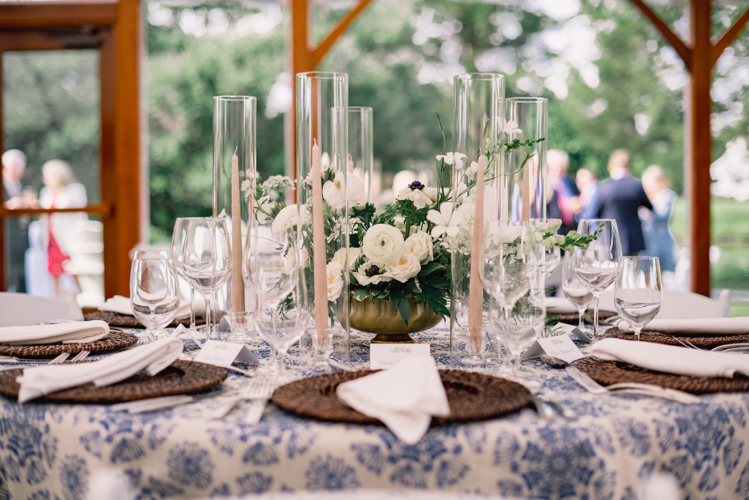 A beautifully set outdoor dining table with a floral centerpiece, surrounded by glassware and place settings under a wooden structure.