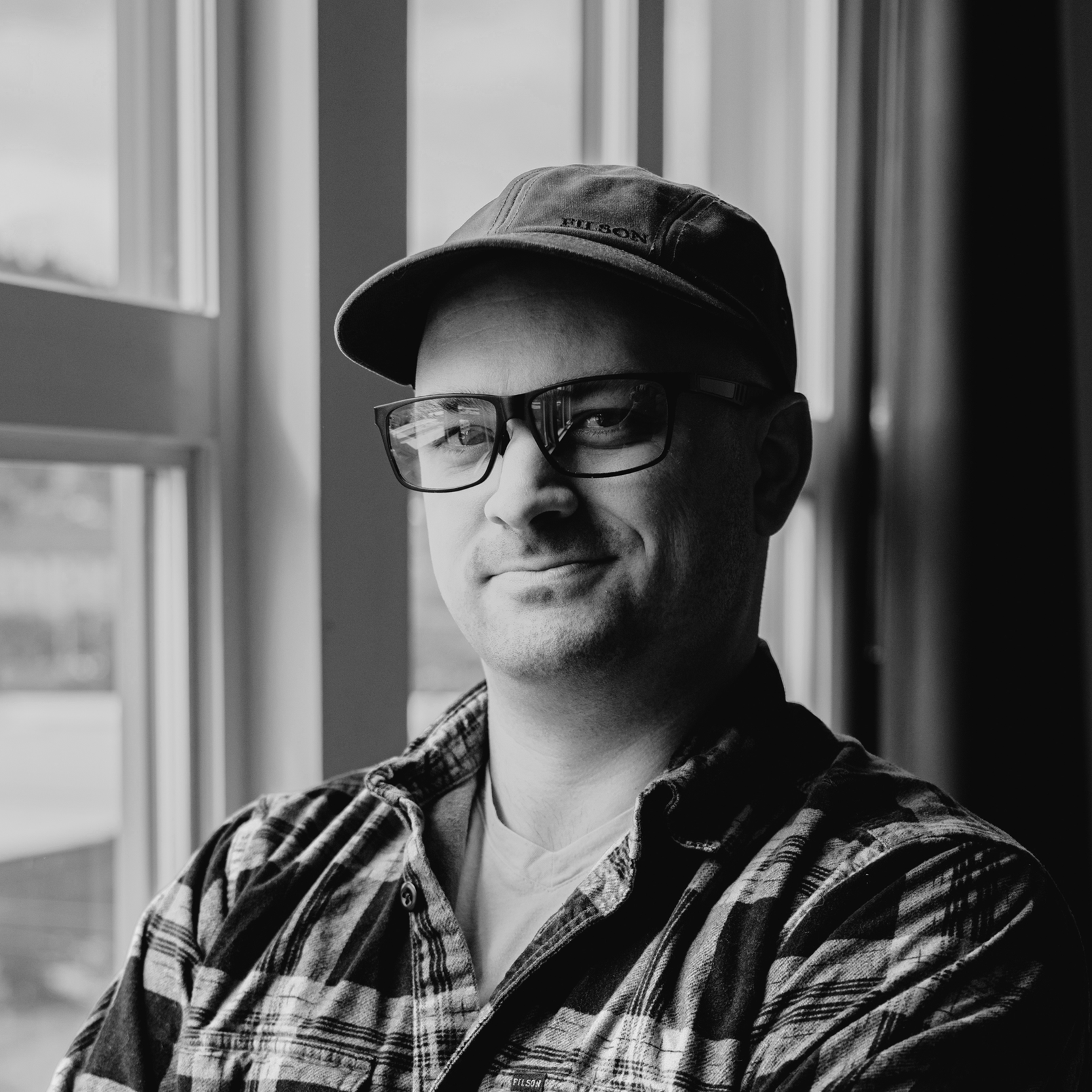 A man in glasses, a cap, and a red and black plaid shirt standing with his arms crossed by a window, looking at the camera with a slight smile.