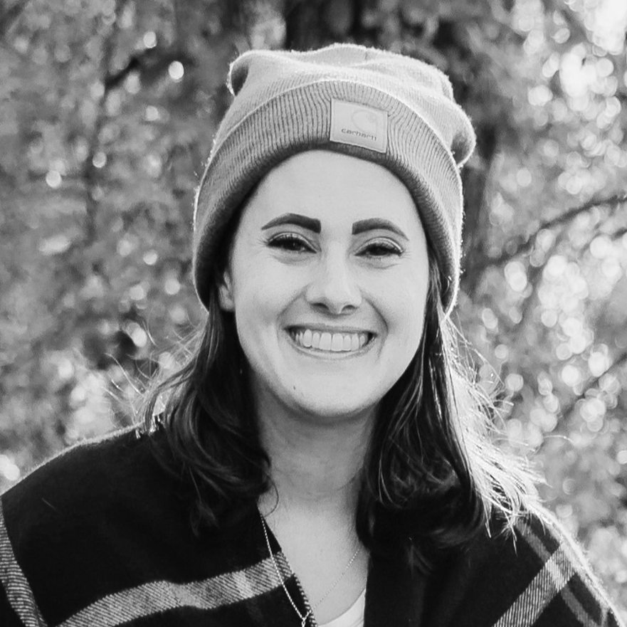 A woman smiling outdoors in fall, wearing a brown Carhartt beanie and a dark plaid jacket with red and white stripes.