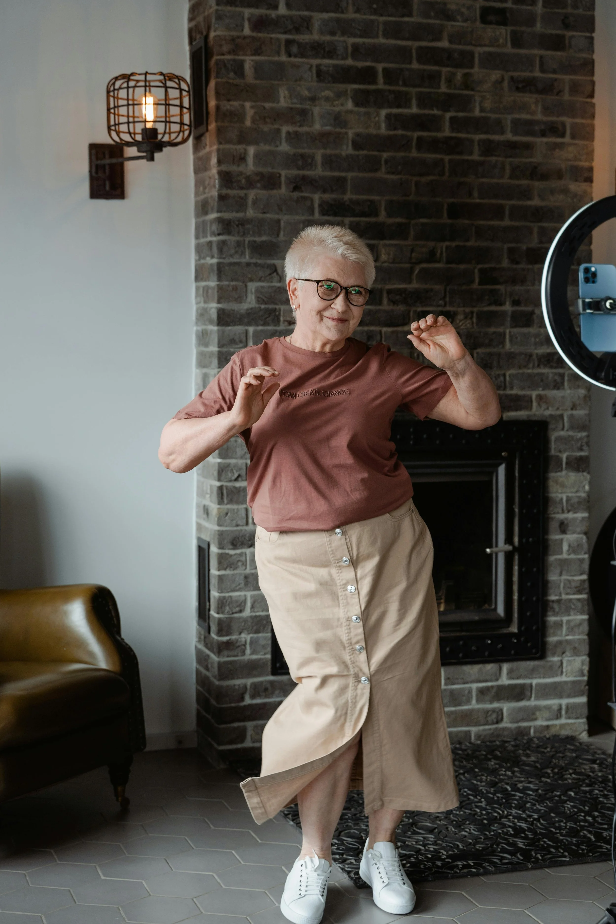 Elderly woman dancing happily indoors, wearing glasses, a brown t-shirt, beige skirt, and white sneakers, near a brick wall and cozy armchair.
