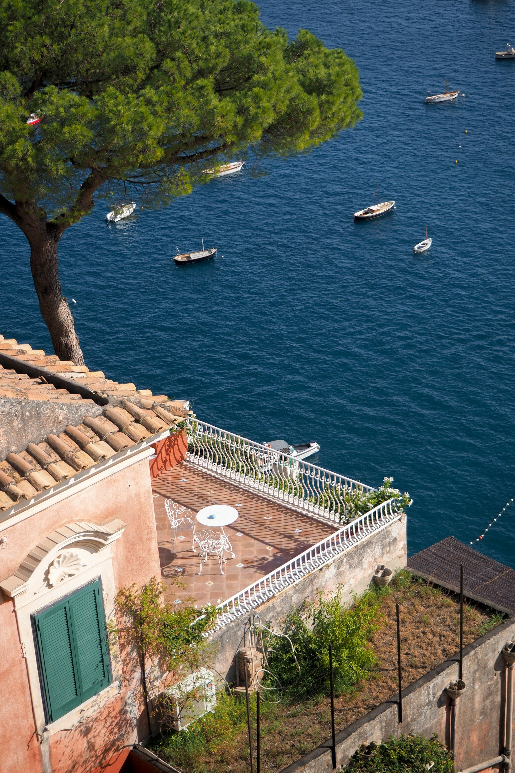 Vue d'une terrasse ensoleillée avec une table, deux chaises et un parasol, face à la mer avec plusieurs voiliers.