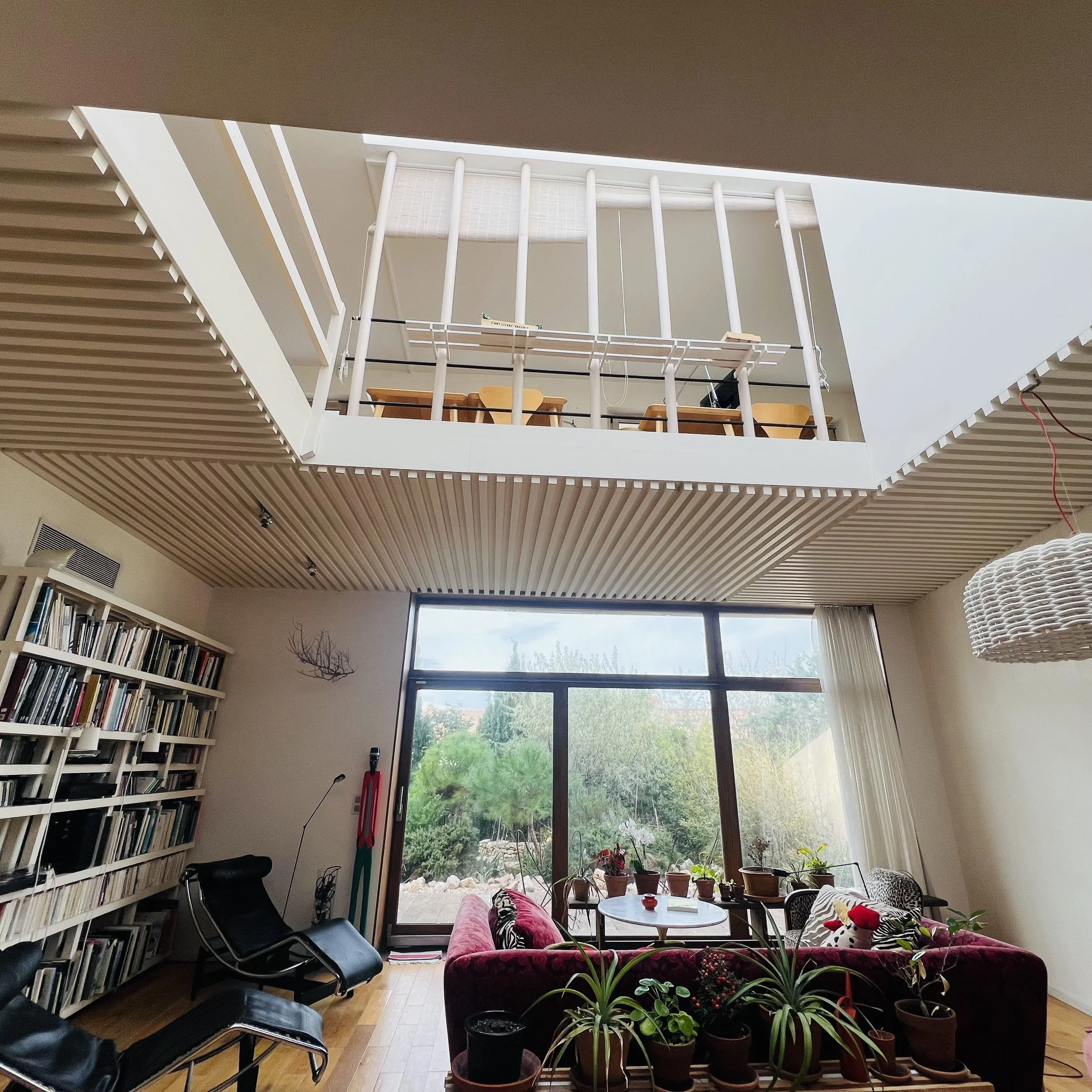 Salon intérieur avec grande fenêtre donnant sur un jardin, plusieurs plantes en pot, canapé rose, tables basses, étagère remplie de livres, chaises, espace lumineux.