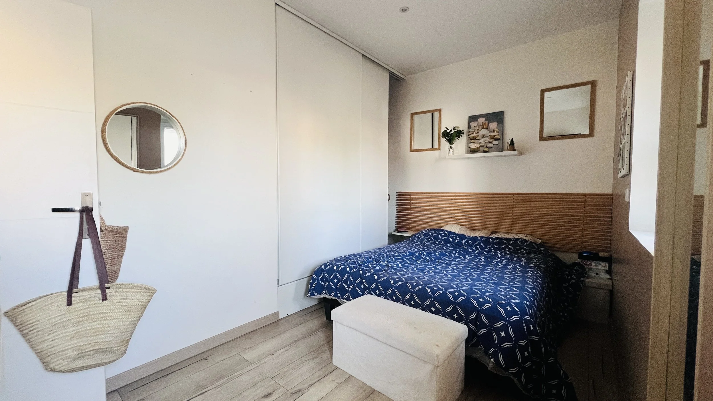 Chambre avec lit double, lit d'appoint et décor simple, miroir carré, cadre rond, tableau, étagère avec plantes et pierres, miroir au mur, porte en bois, sol en bois clair.