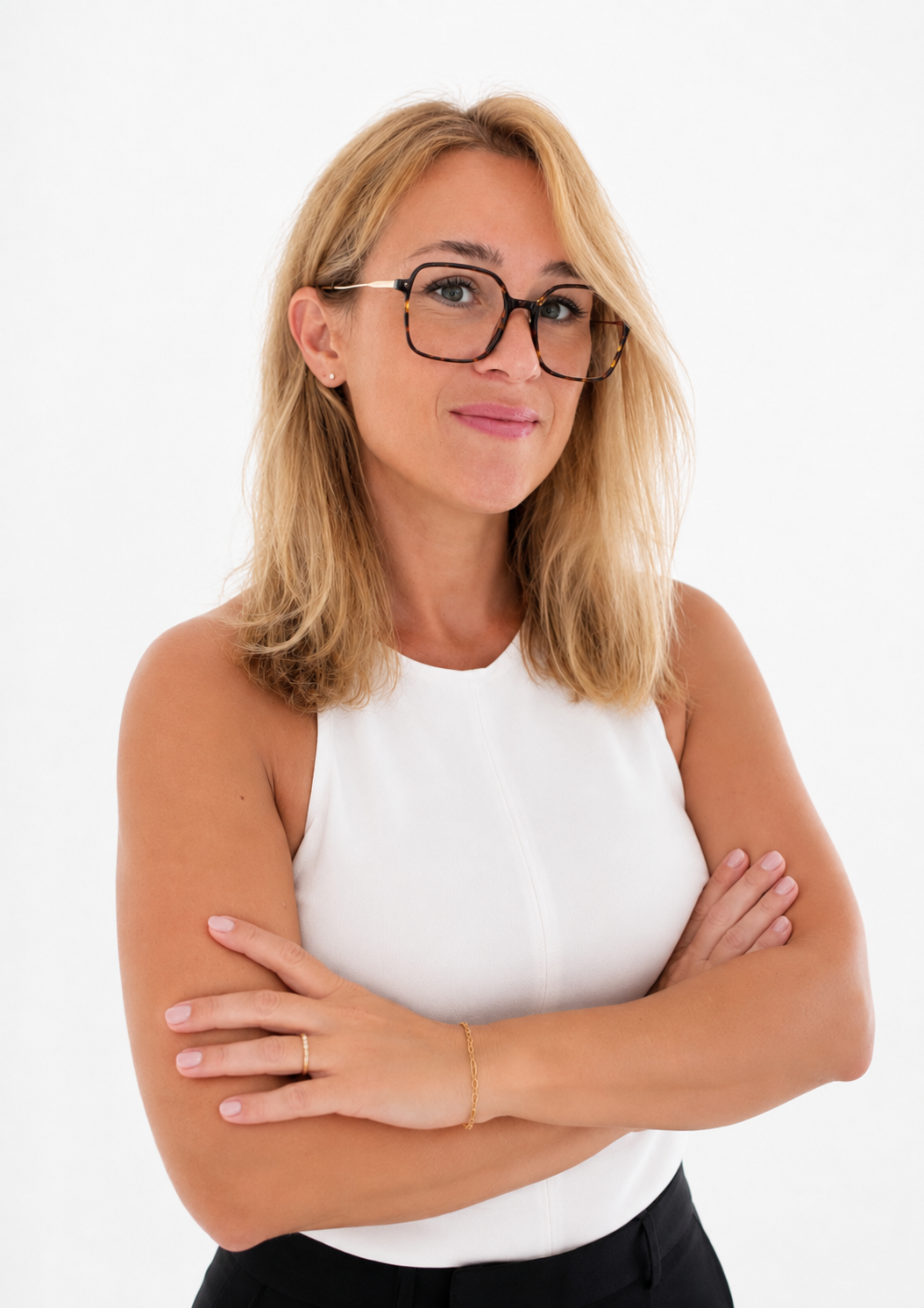 Une femme blonde portant des lunettes, une robe blanche et un bracelet, croisant les bras, contre un fond blanc.