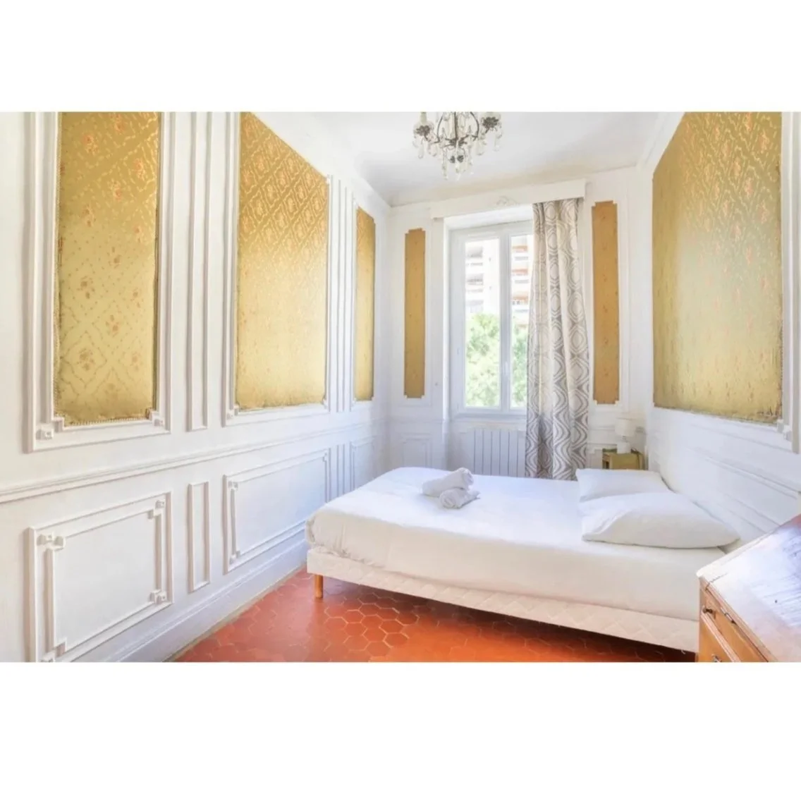 Chambre avec un lit, un aïlou et des murs décorés de panneaux dorés, fenêtres laissant entrer la lumière, rideaux crème, plafond avec lustre en cristal.