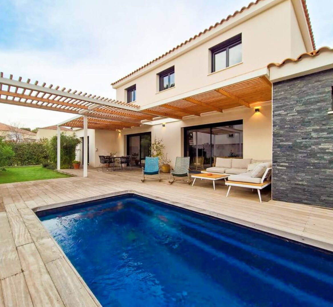 Une maison moderne avec un patio en bois, un lit de piscine bleu, un espace salon avec un canapé beige et des chaises, et une pergola en bois.
