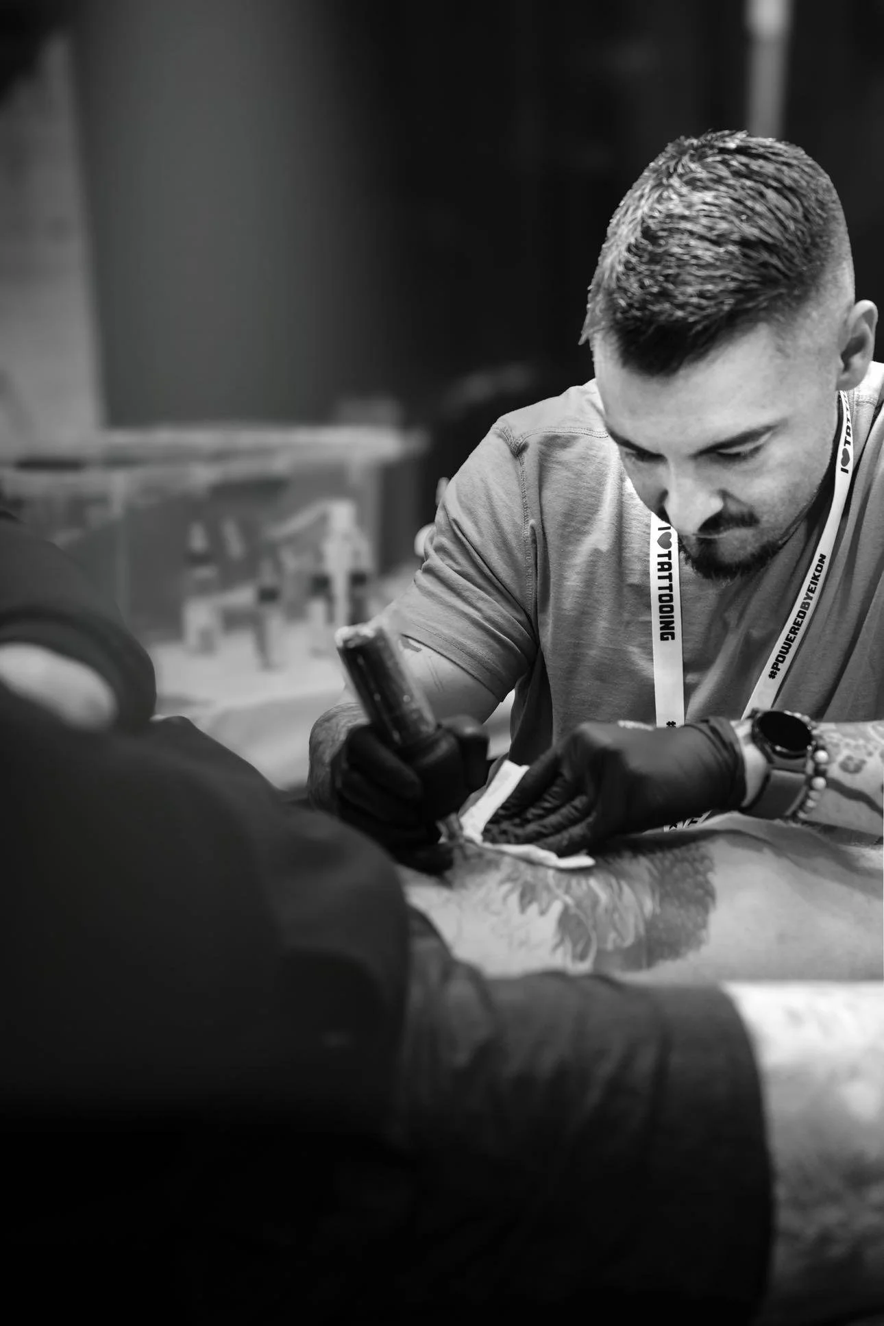 A man with short, styled hair and a beard, wearing gloves, is sitting at a table, focused on his phone. He is wearing a lanyard and a wristwatch. The background appears to be a workshop or gym with equipment and bottles visible.