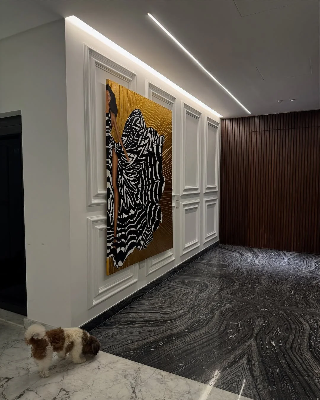 Interior of a modern room with white and wooden paneled walls, black and white marble flooring, and a portrait of a woman in a black and white striped dress hanging on the wall. There is a small dog near the corner of the white marble floor.
