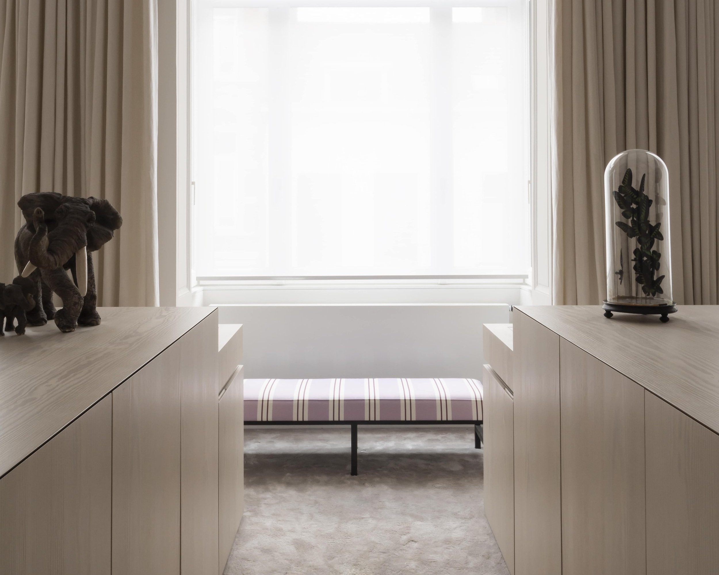A minimalist room with light wood cabinets on both sides, a padded bench with striped upholstery under a large window with white curtains, and decorative items including elephant figurines and a glass dome with plants.