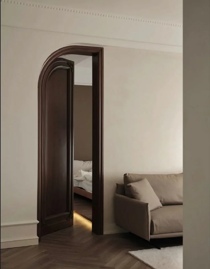 A view of a living room showing an open wooden door with rounded edges leading to a bedroom, with a beige sofa with two cushions on the right side and a small white rug on the wooden floor.