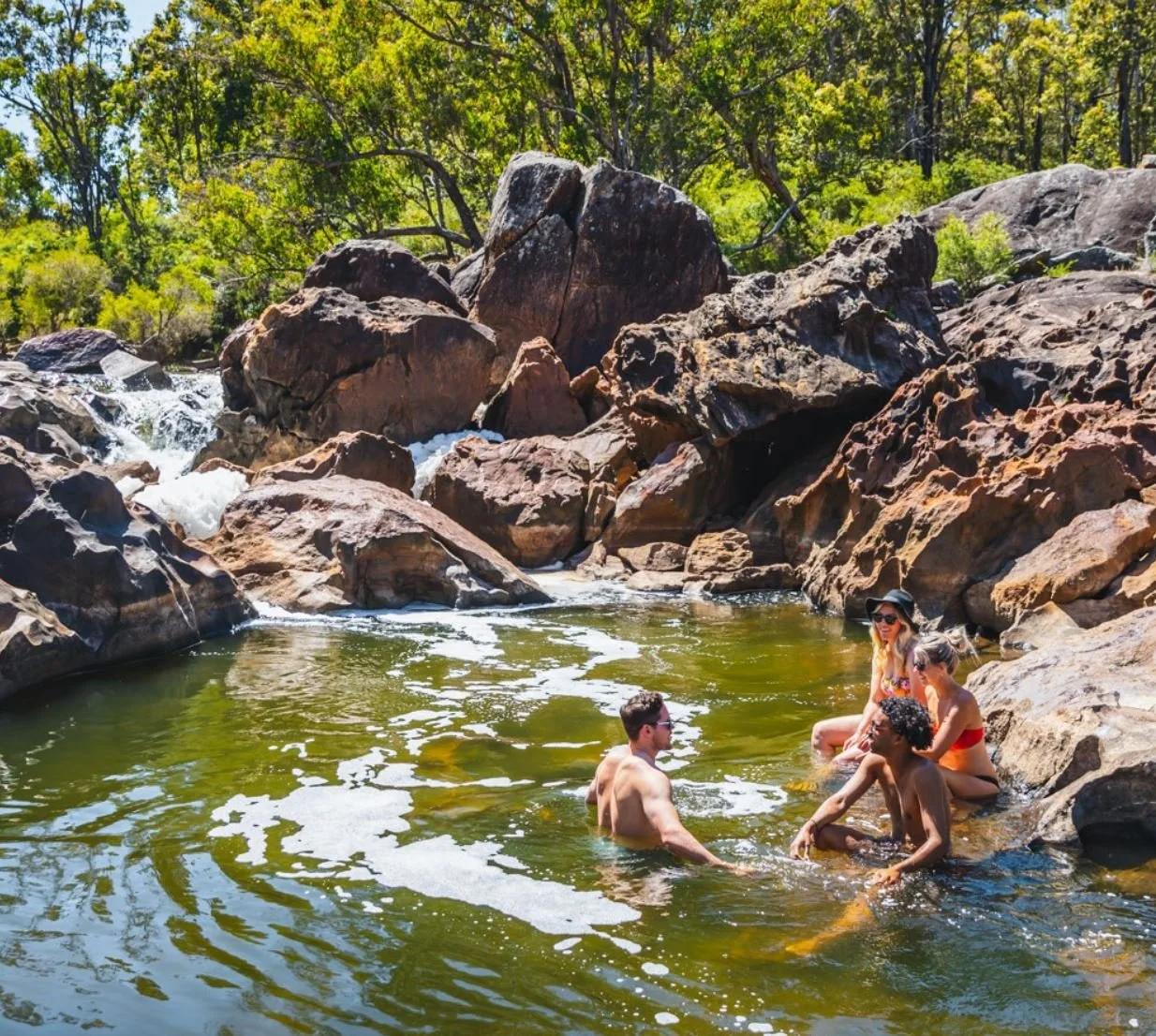 Four people sitting and swimming in a shallow river with large rocks, trees, and a small waterfall in the background on a sunny day.