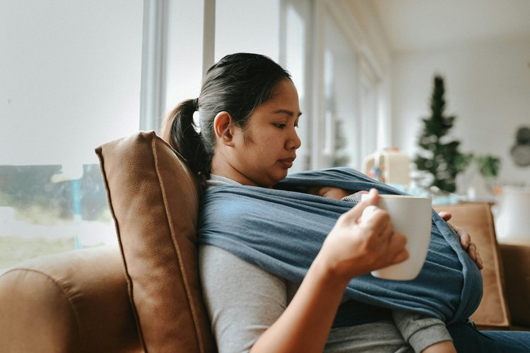 A mother sitting on the couch with a warm drink and her baby in a carrier