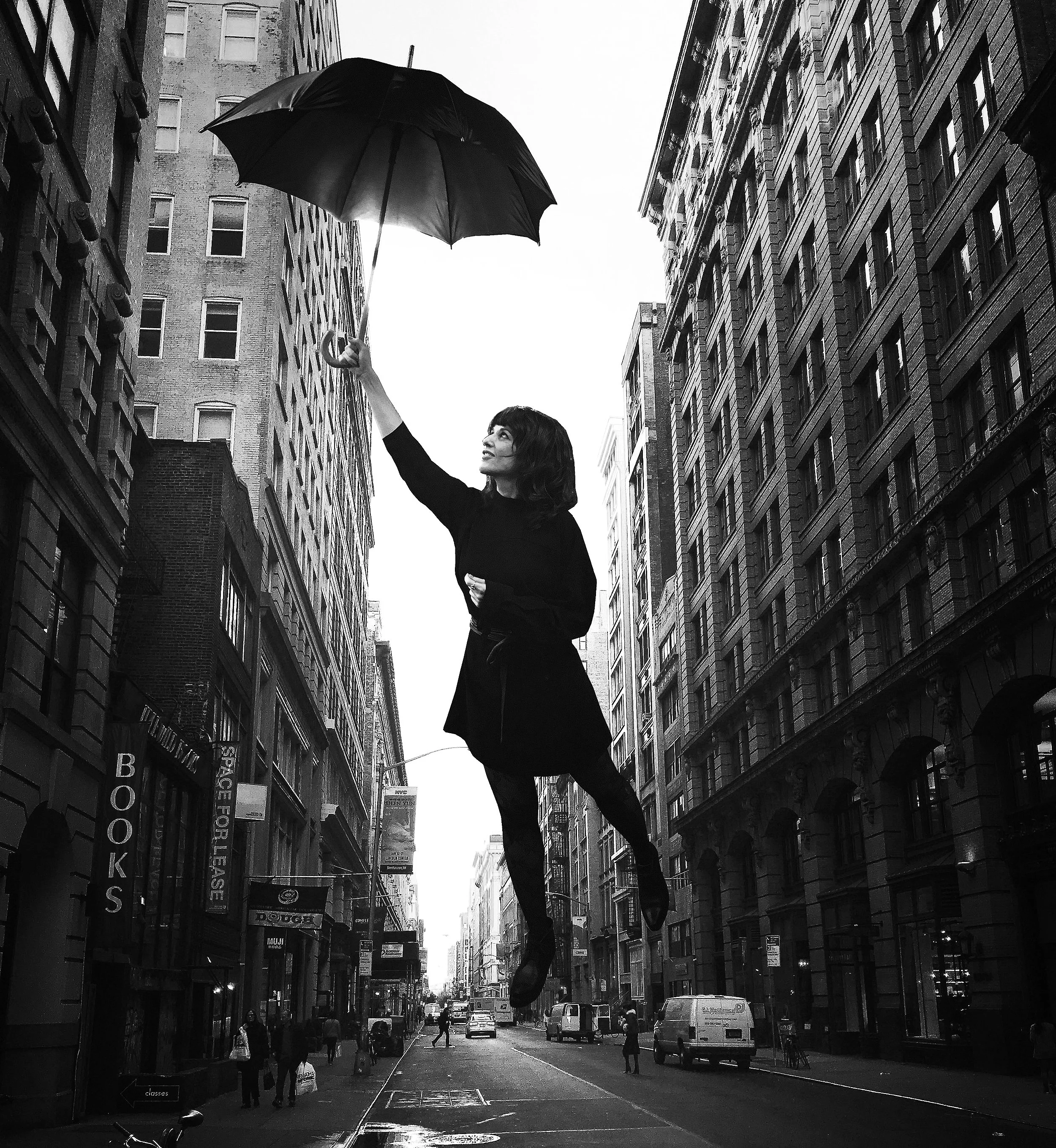 A woman in a black outfit appears to be floating in the air while holding an umbrella on a city street with tall buildings.