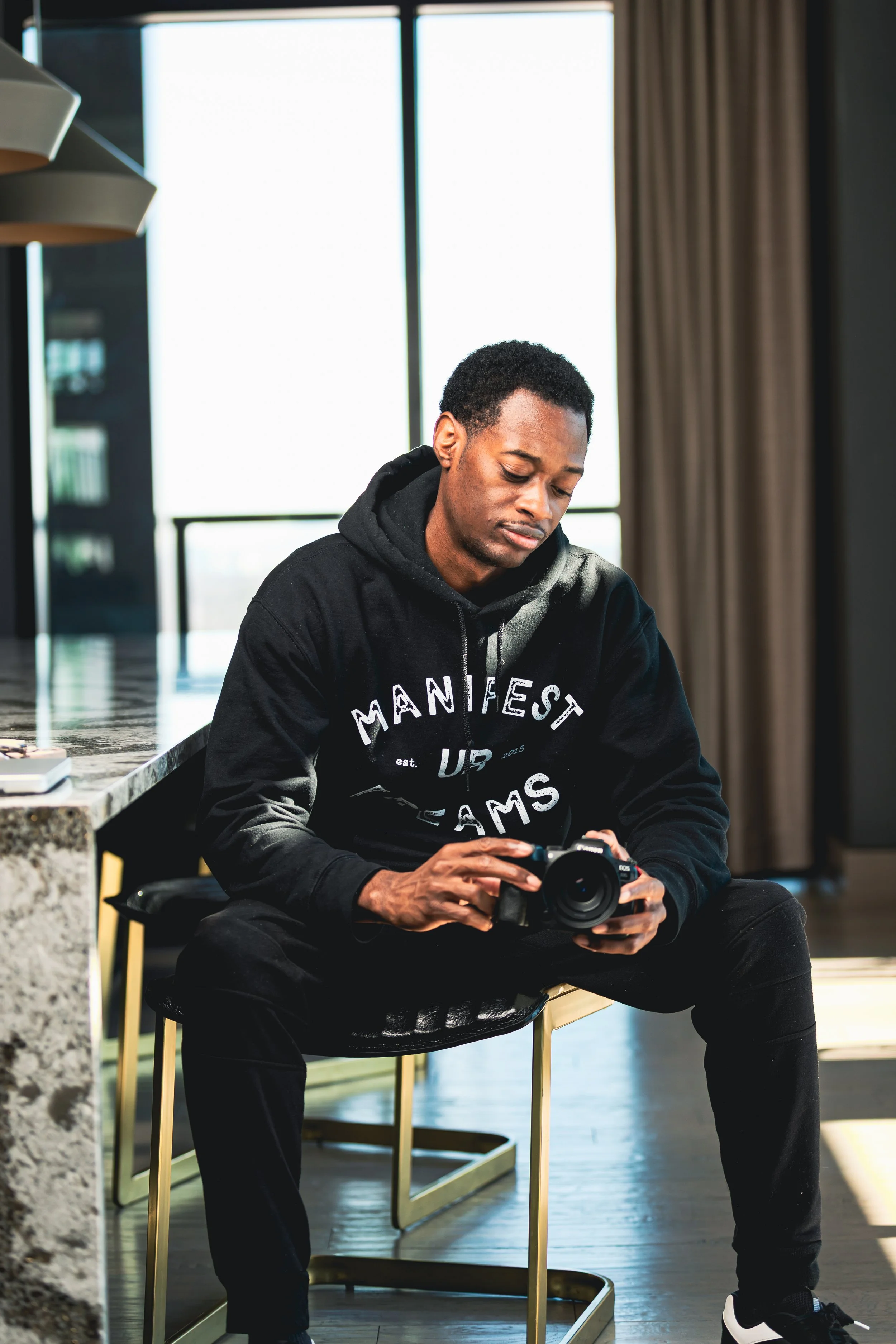 A young man sitting on a modern chair in a bright room, holding a camera and looking down at it with a focused expression.