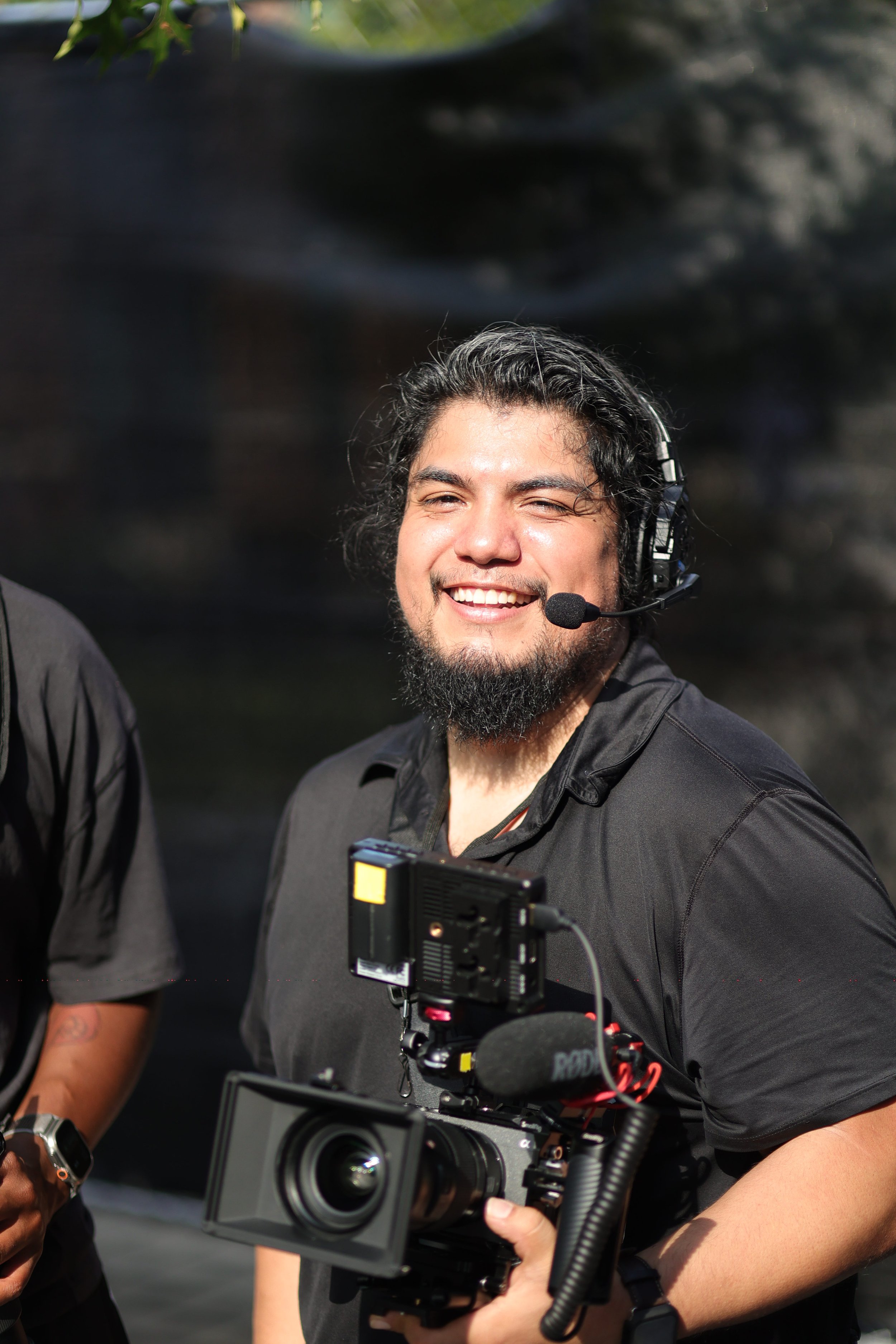 A man with long dark hair, a beard, smiling, wearing a black shirt, with a camera and a microphone, standing outdoors.
