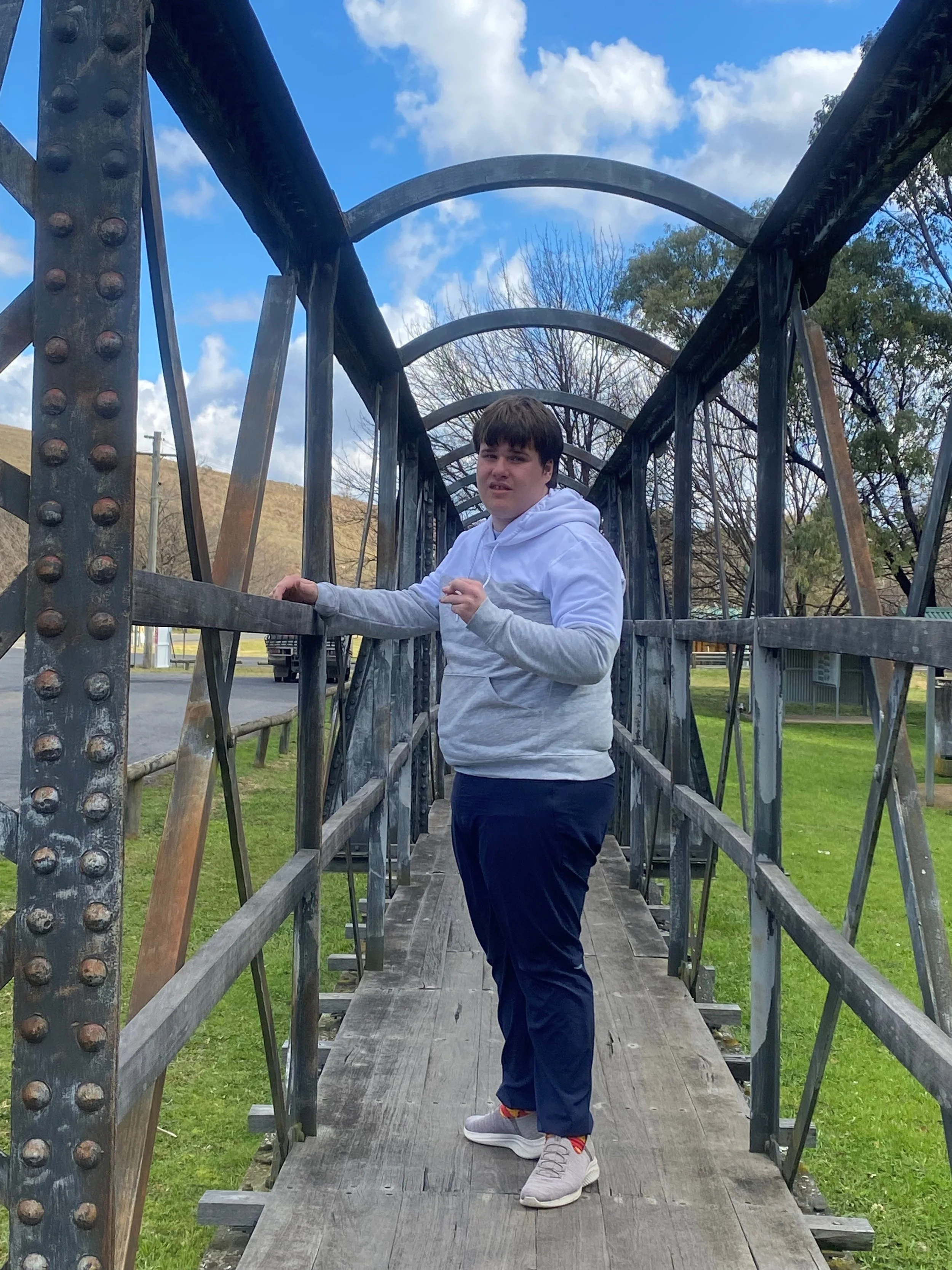 A young man wearing a white hoodie, navy blue pants, and sneakers stands on a wooden and metal footbridge outdoors. The sky is partly cloudy, and there are trees and grassy areas in the background.