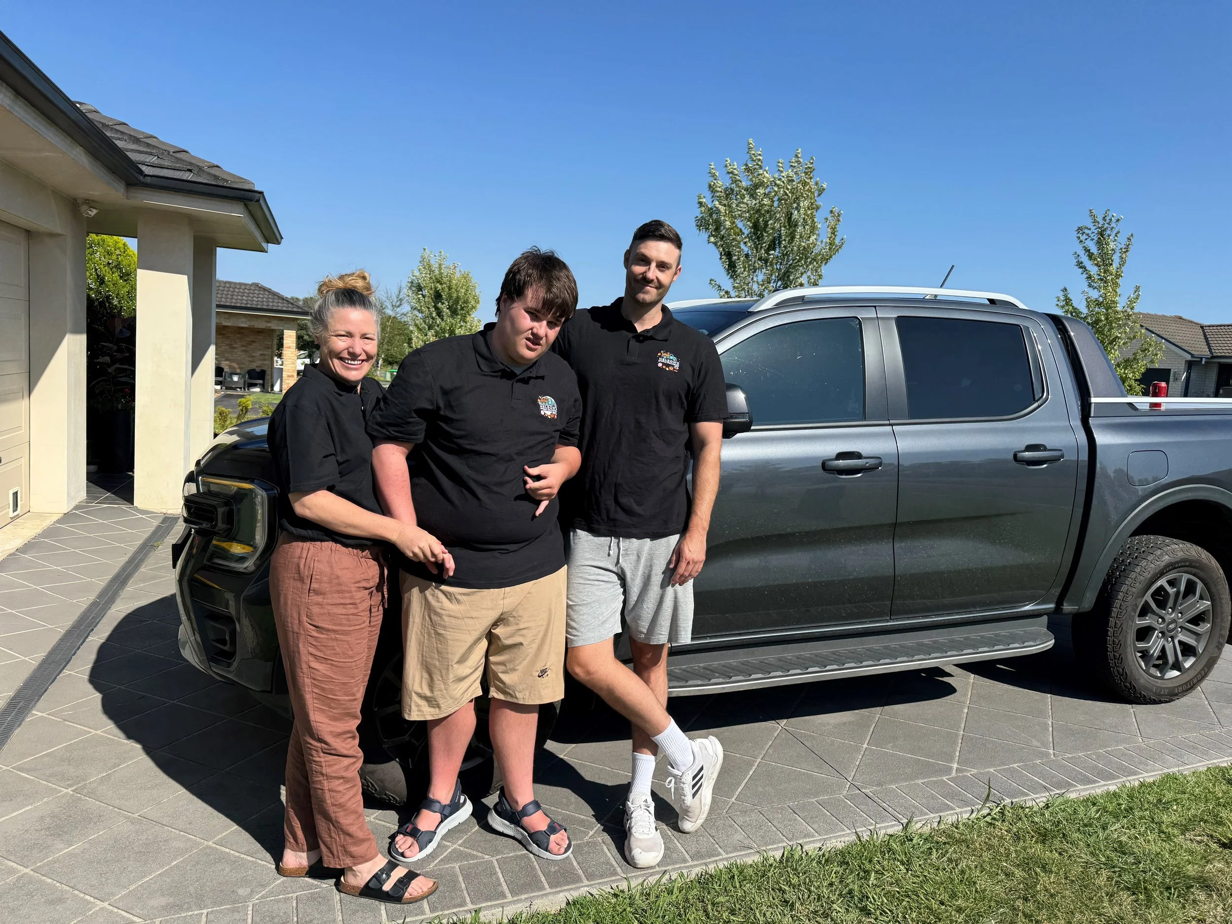 Three smiling people standing in front of a silver pickup truck in a residential neighborhood on a sunny day. There are houses and trees in the background. Fresh vegetable and fruit box available for delivery in Orange NSW