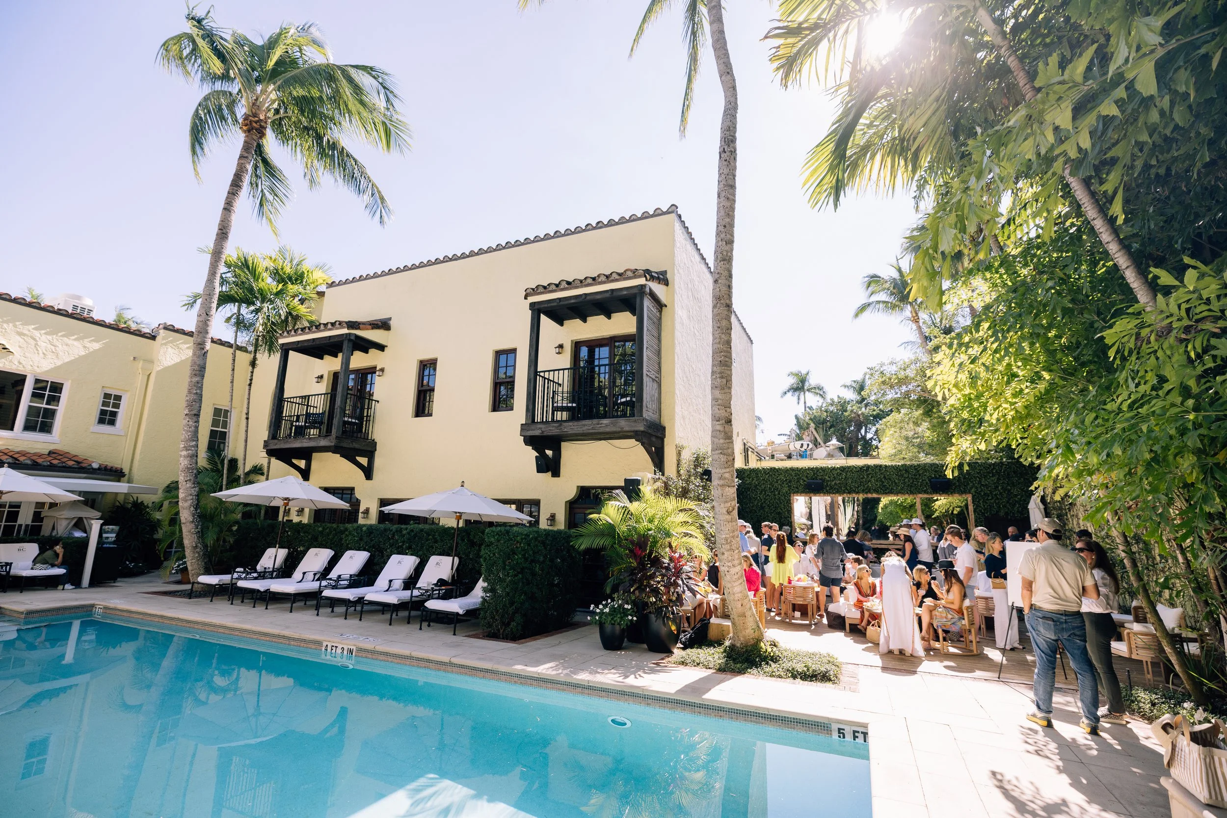 A sunny outdoor scene featuring a swimming pool surrounded by lounge chairs under umbrellas, with a yellow Mediterranean-style building in the background and palm trees. A group of people is gathered on the right, possibly at a social event.