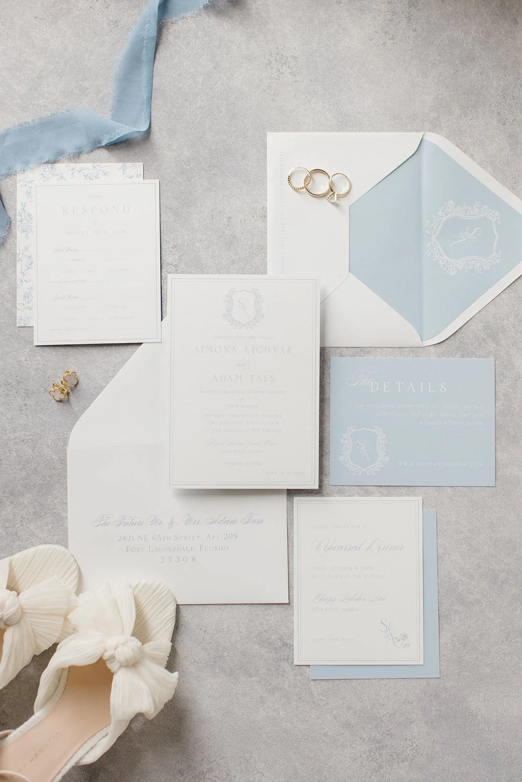 Flat lay of wedding invitation suite with blue and white theme, including RSVP card, details card, envelopes, rings, and bow-accented shoes on a gray background.