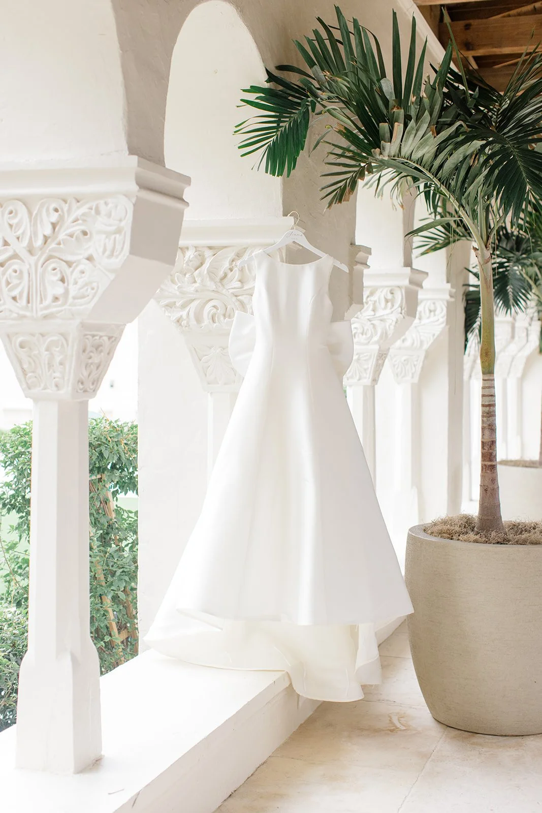 Elegant white wedding dress hanging on a hanger against a white decorative wall with archways and a potted palm plant.