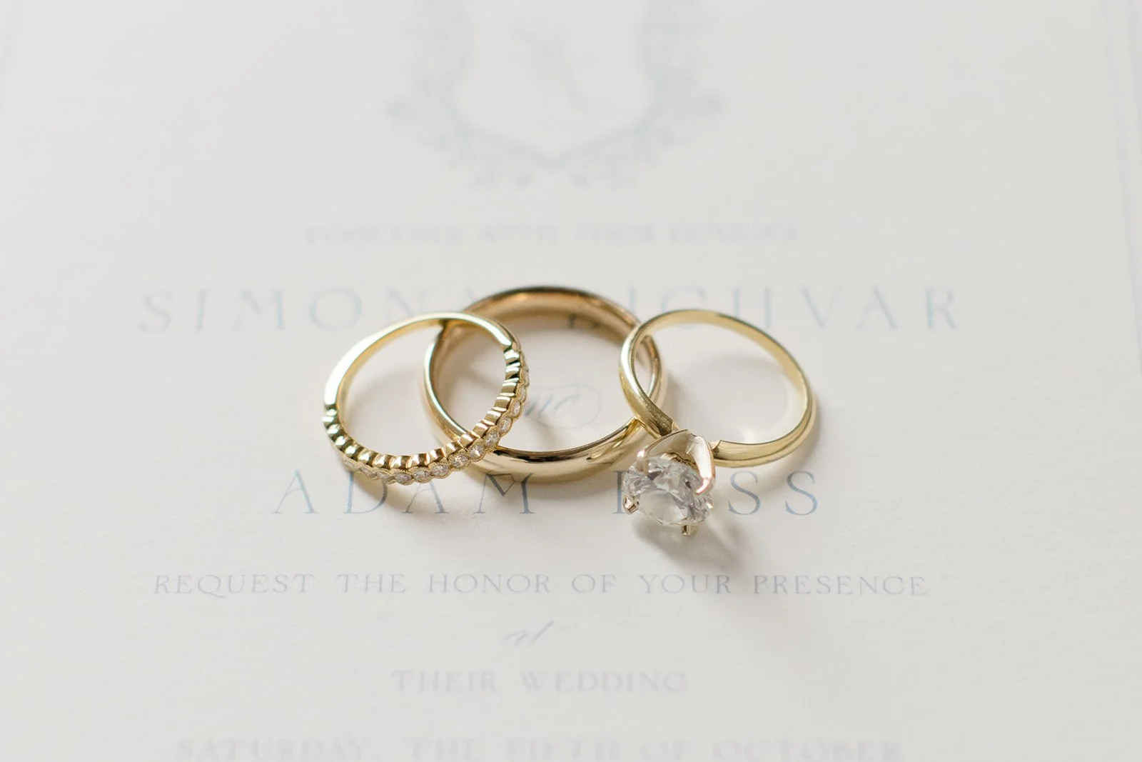 Three gold rings, including a diamond solitaire engagement ring and a wedding band, on a wedding invitation.