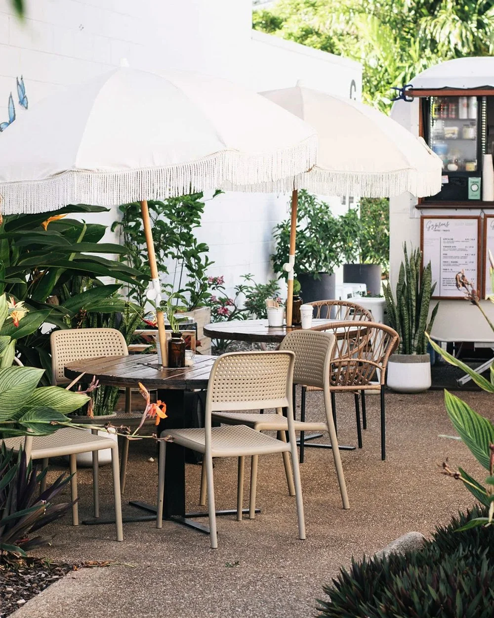 Outdoor seating area with tables, beige chairs, two white umbrellas, and surrounding greenery at a cafe or patio.