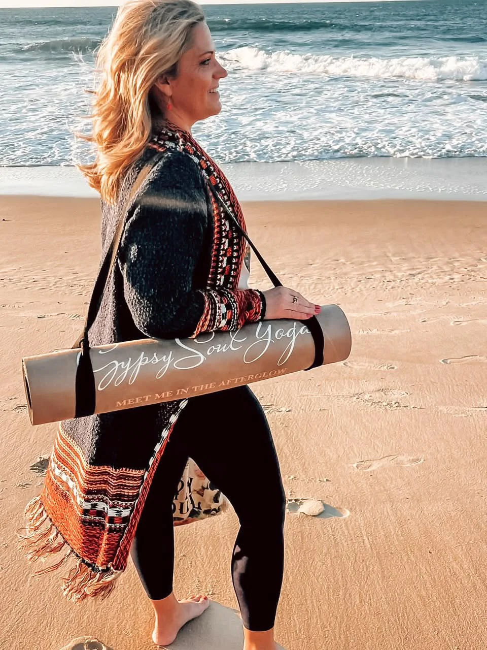 Woman walking barefoot on a sandy beach while carrying a yoga mat that reads "Gypsy Soul Yoga" and "Meet me in the afterglow," with the ocean waves in the background.