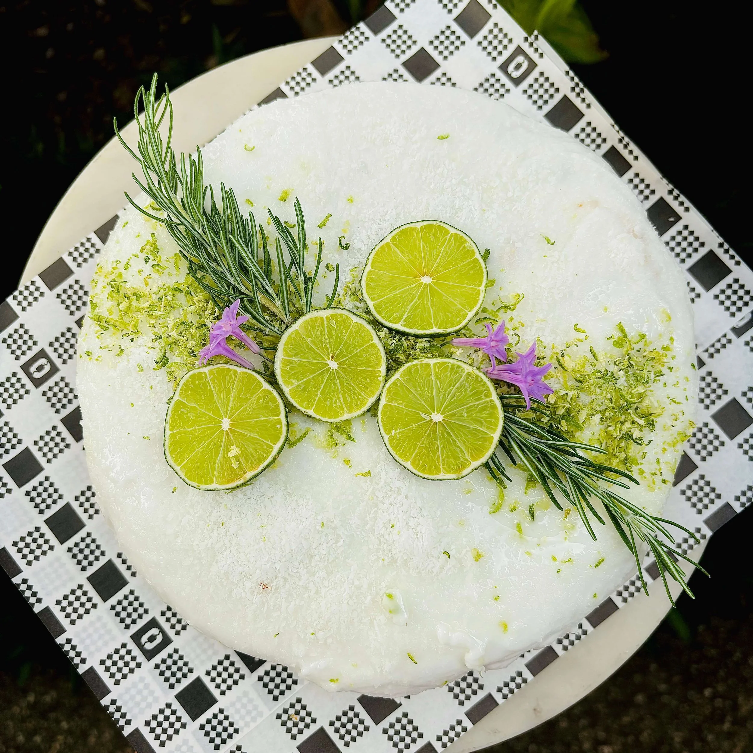 Close-up of a white cake decorated with lime slices, purple flowers, and sprigs of rosemary on a patterned paper doily.