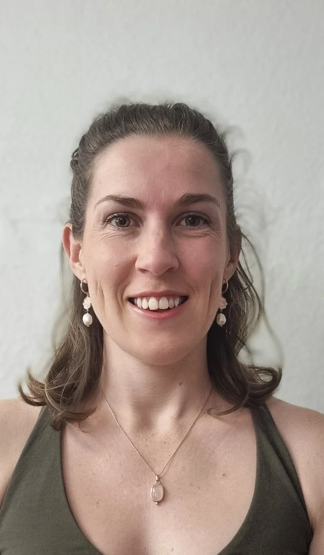 Close-up of a smiling woman with shoulder-length brown hair, wearing earrings and a necklace, against a plain light background.