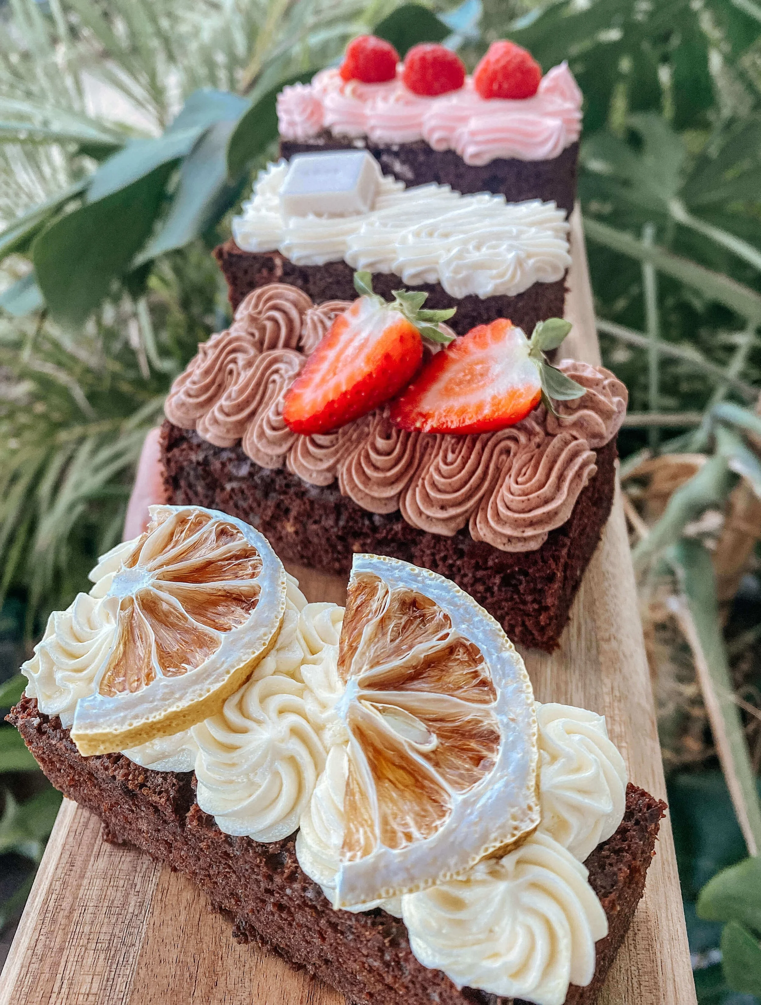 Three slices of chocolate cake decorated with strawberries, whipped cream, lemon slices, and orange slices, placed on a wooden board with green plants in the background.
