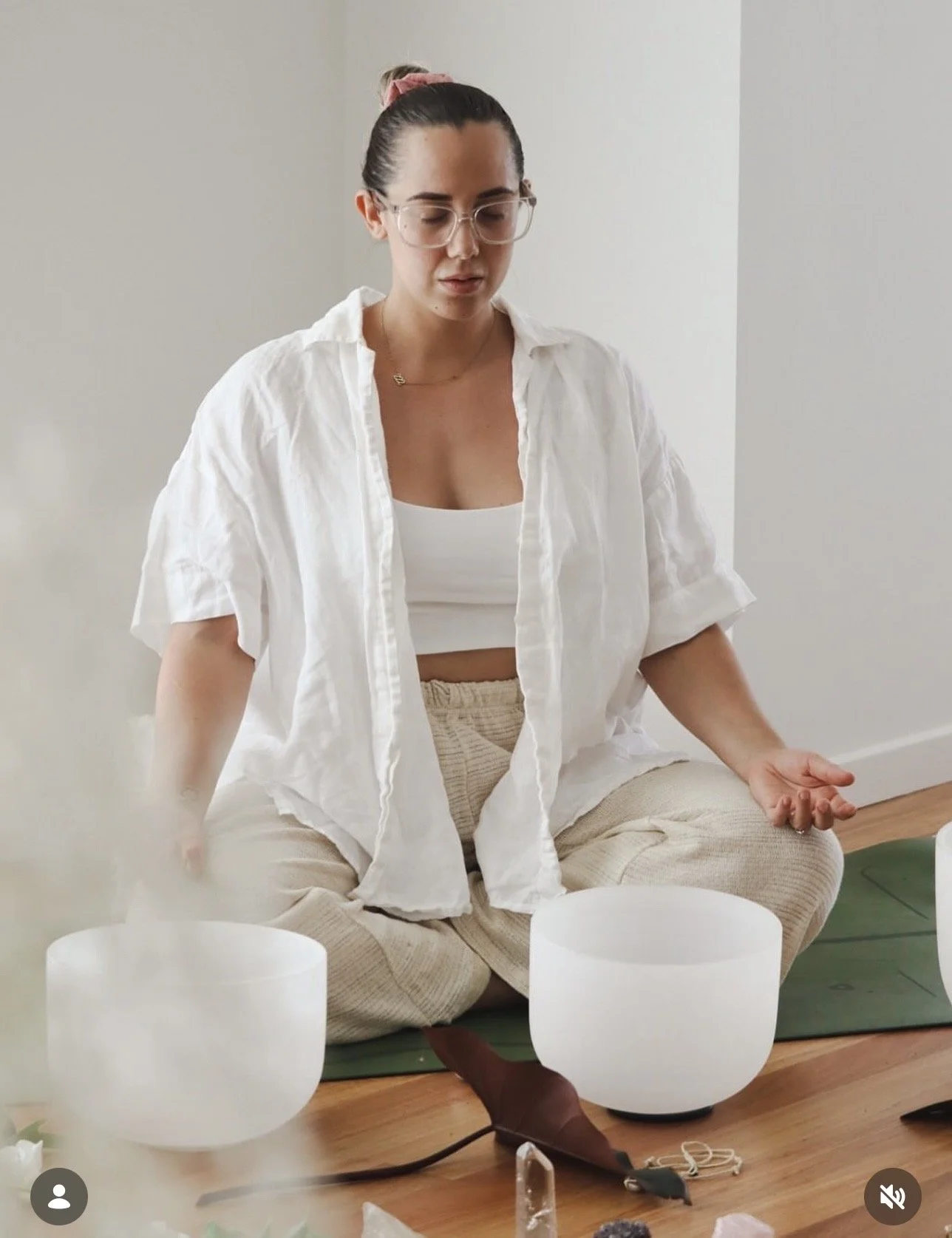A woman sitting cross-legged on a cushion on the floor, with her eyes closed, wearing glasses, a white top, and beige pants. She appears to be meditating or practicing mindfulness. In front of her, there are several white singing bowls on a green mat.
