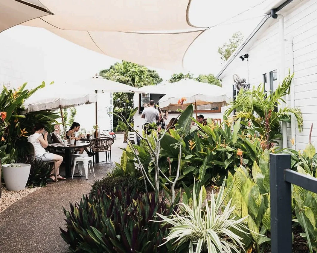Outdoor patio of a cafe or restaurant with large white umbrellas, lush green plants, and several patrons enjoying meals or drinks.