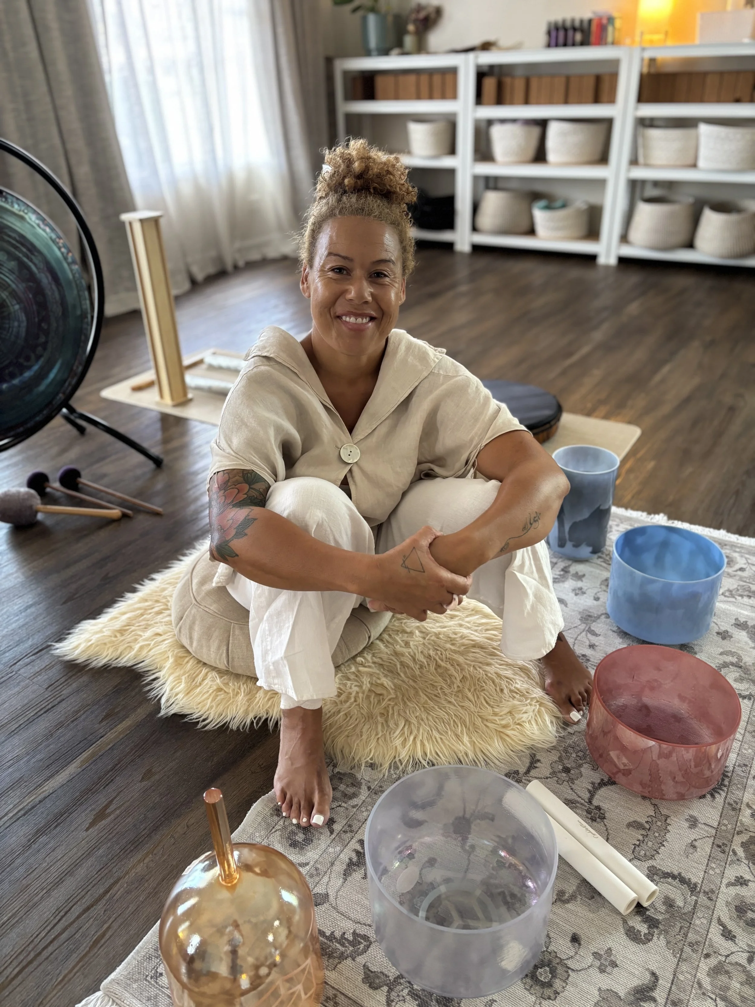 A woman with curly blonde hair sitting on a fluffy beige rug. She appears to be in a room with white shelves filled with baskets and books. She is smiling and looking at the camera, with a tattoo on her left arm. The room has a wooden floor, and there are colorful singing bowls and a metallic sphere with a straw in the foreground.