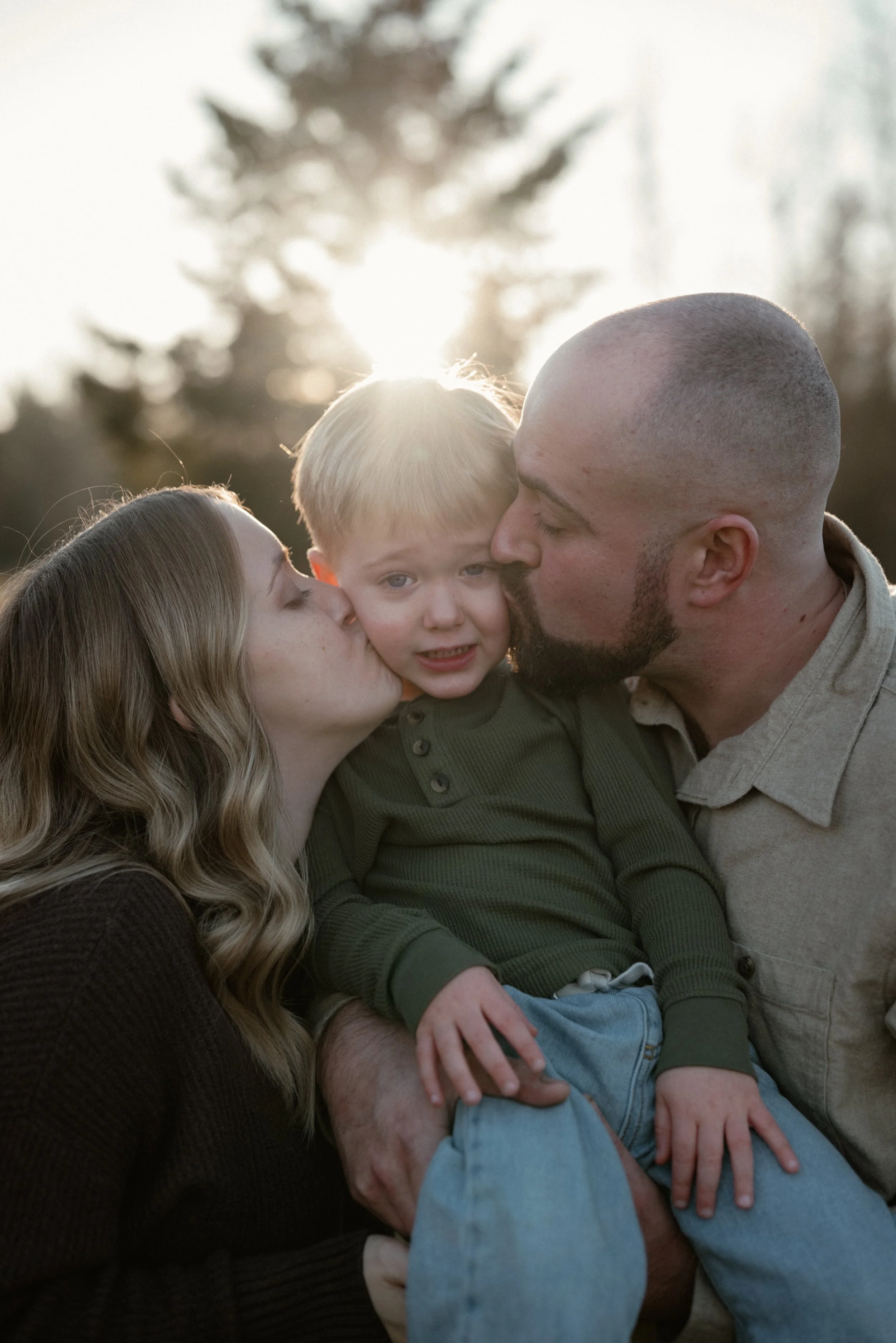 A family of three, a woman, a man, and a young boy, outdoors during sunset. The woman and man are kissing the boy on his cheeks, and the boy is looking at the camera with a slightly nervous expression. Trees are blurred in the background with sunlight shining behind them.
