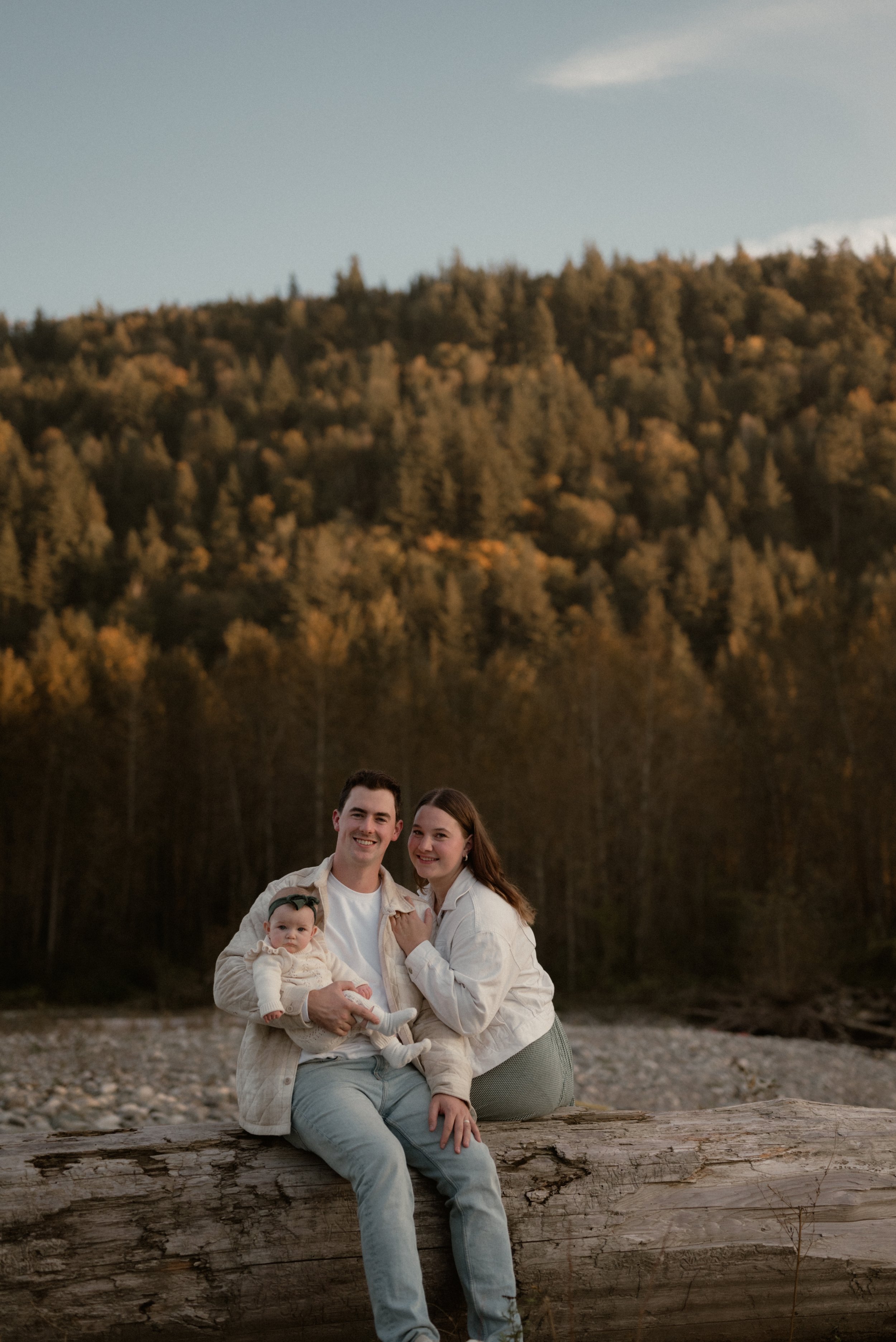 A family of three sitting on a large log outdoors with a forested hillside in the background, during golden hour.