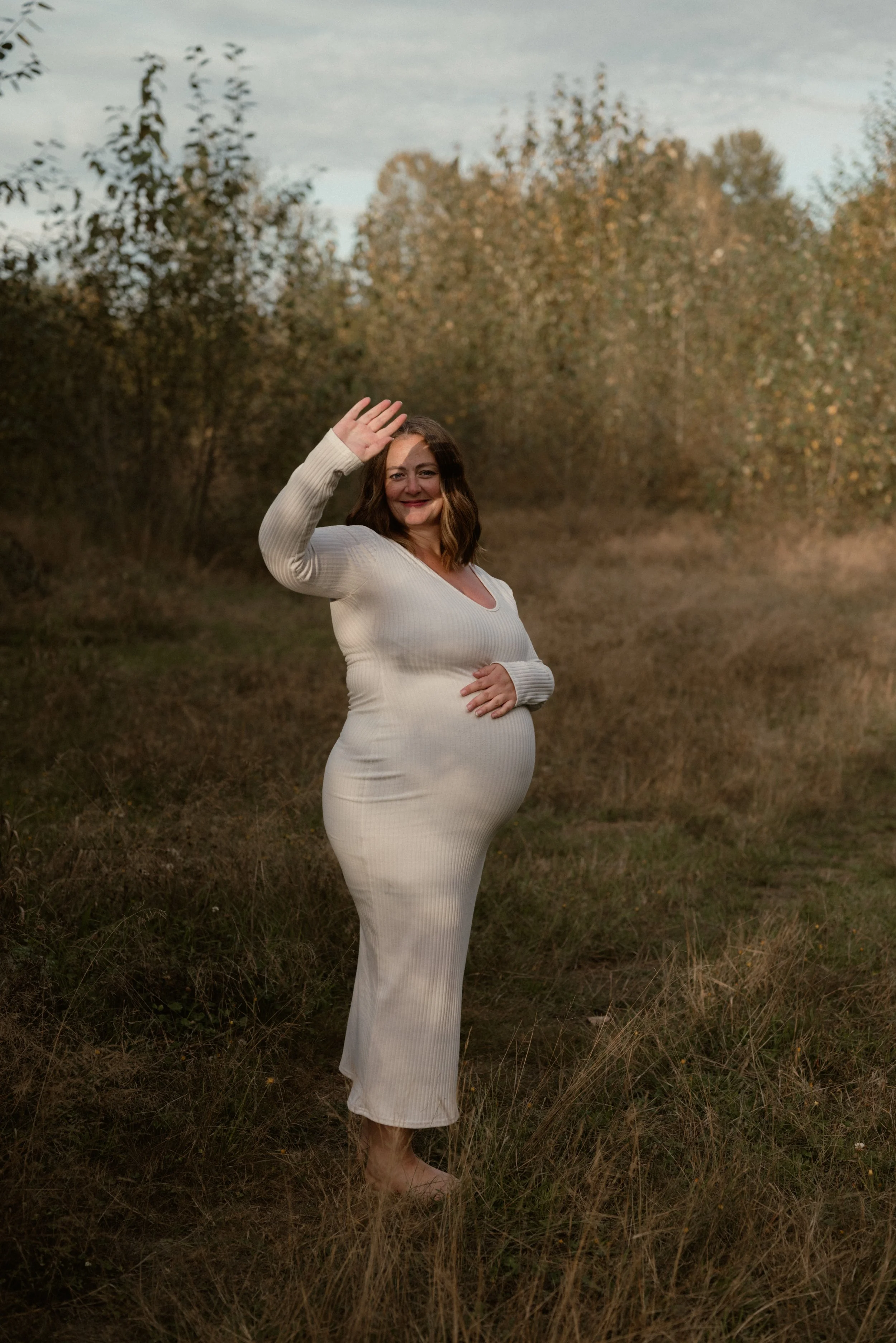 A pregnant woman in a white long-sleeve dress standing outdoors in a grassy field with trees in the background, smiling and waving.