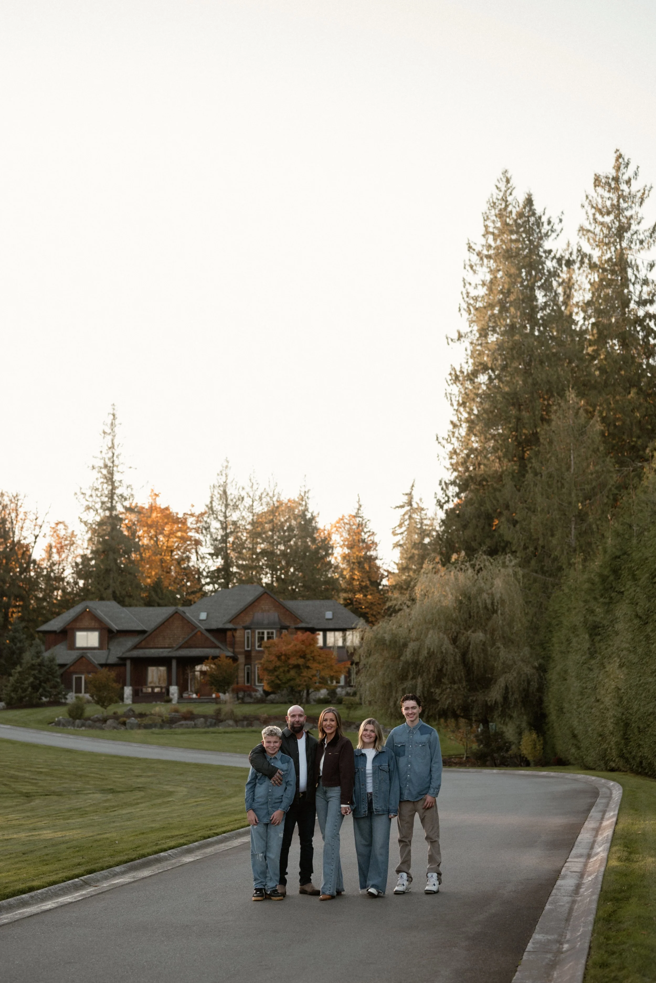 A family of five standing on a driveway in front of a large house with trees in the background during autumn.
